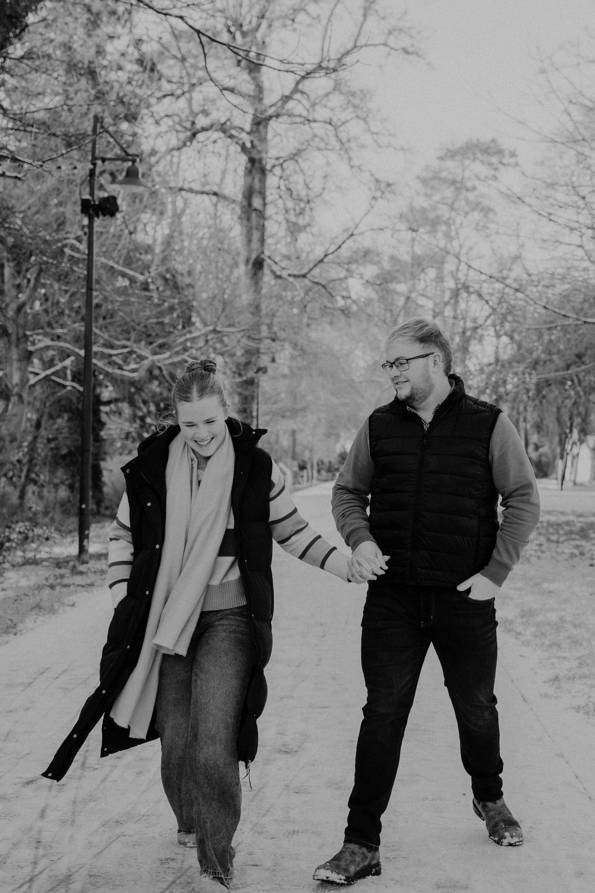 A black and white photo of a smiling young woman and man walking hand in hand outdoors on a sidewalk in a park or wooded area, with leafless trees and a lamppost in the background.