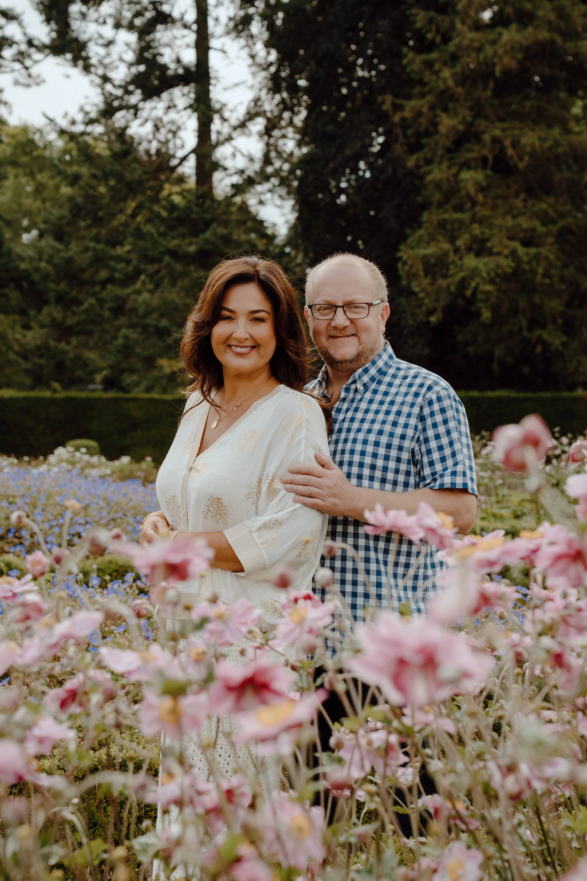 A smiling woman and man standing together in a colorful flower garden with trees in the background.