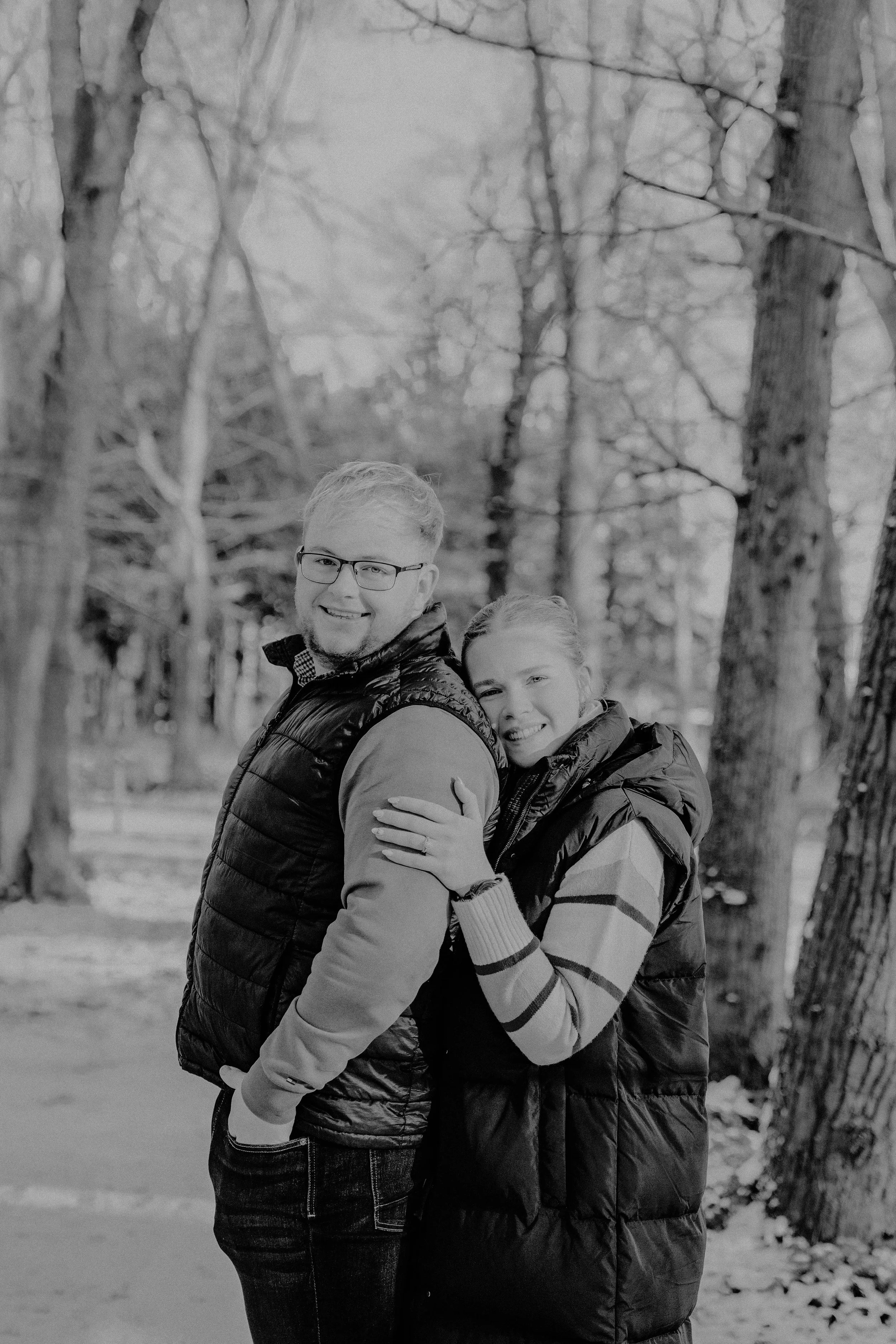 A smiling man and woman standing outdoors in a wooded area, hugging each other, wearing warm jackets.