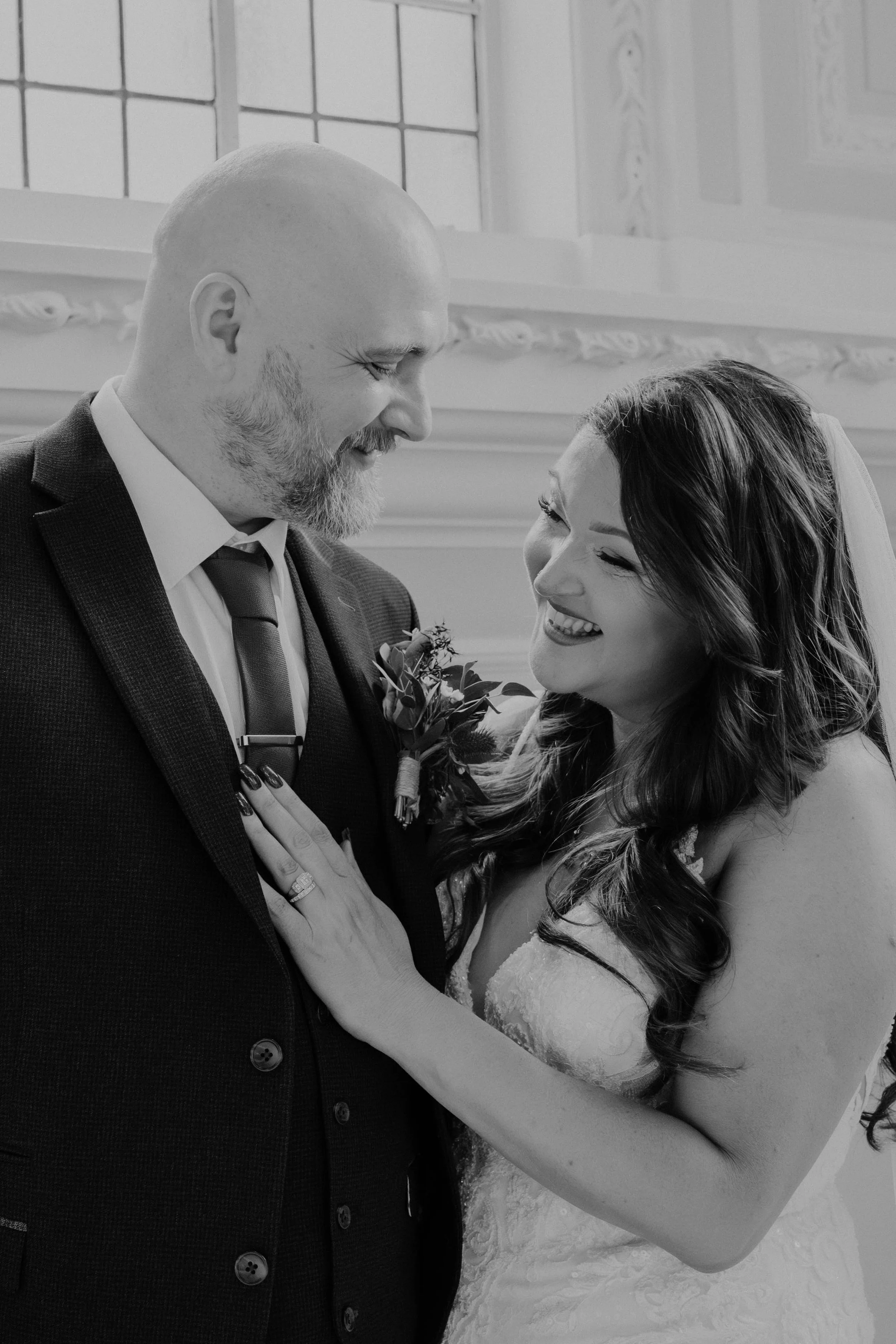 A black-and-white photograph of a bride and groom smiling and looking into each other's eyes during their wedding. The groom has a beard, is wearing a suit with a boutonniere, and the bride has long, wavy hair and a wedding dress with lace details.
