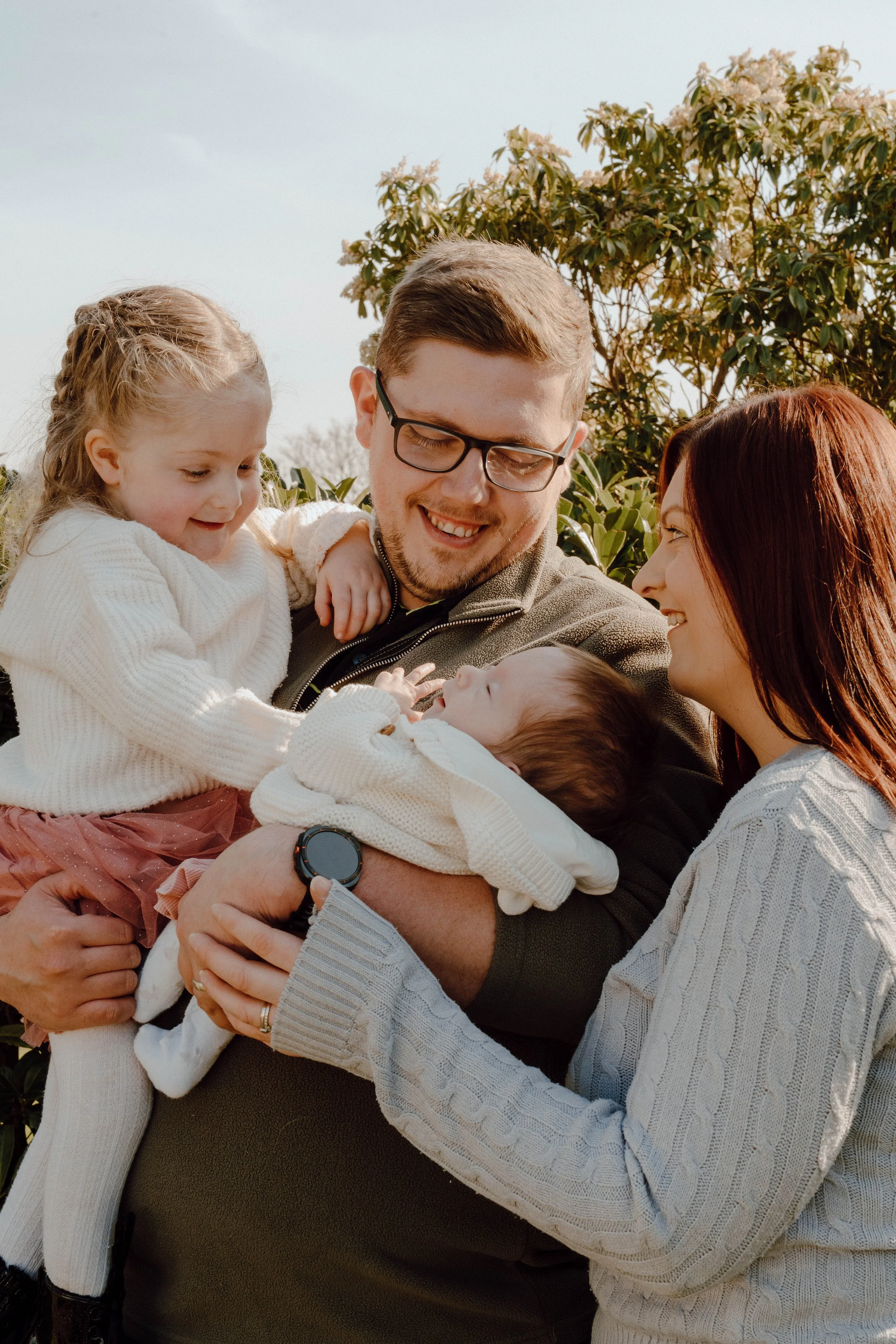Family of four outdoors during daytime, with a man holding a newborn baby, a woman looking at them, and a young girl touching the baby, all smiling.