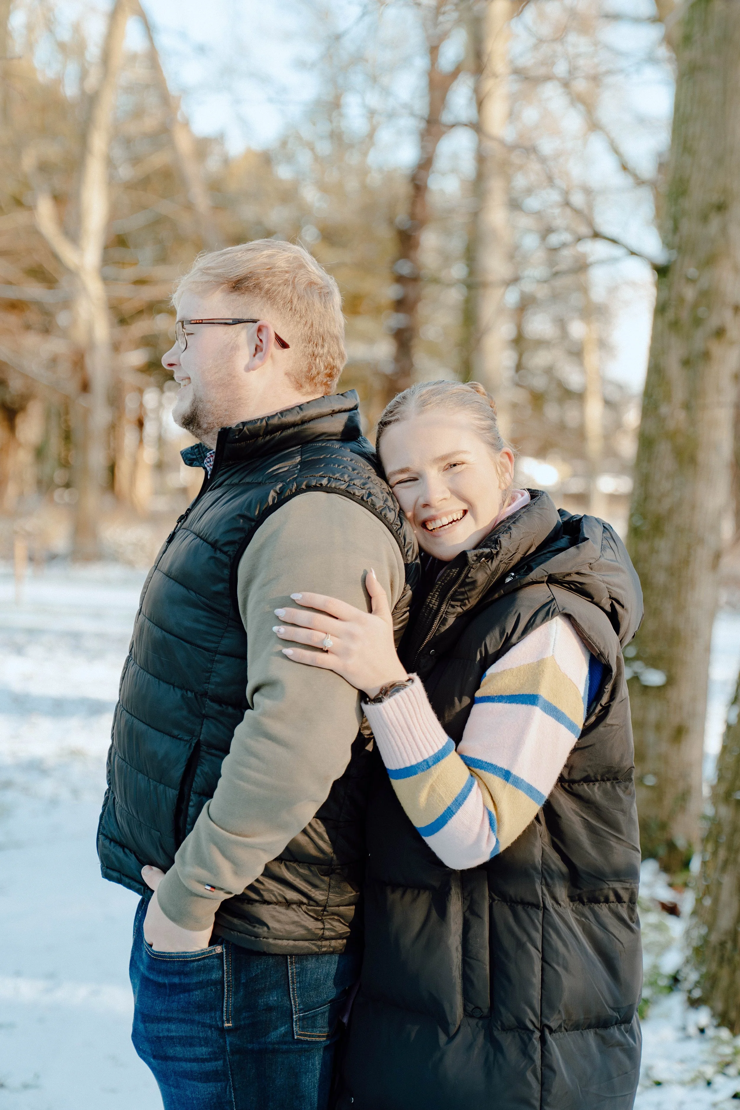 A smiling woman hugging a man from behind outdoors in a snowy park, with trees in the background.