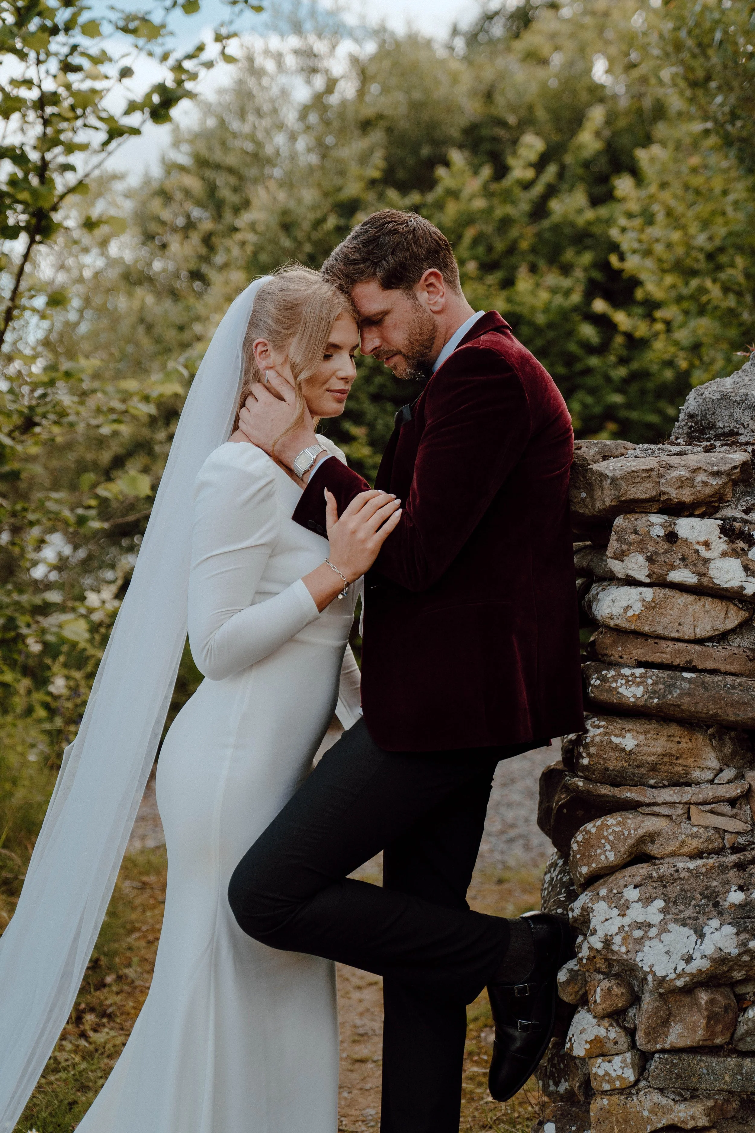 A bride and groom share an intimate moment outdoors, leaning against a stone wall surrounded by green trees.