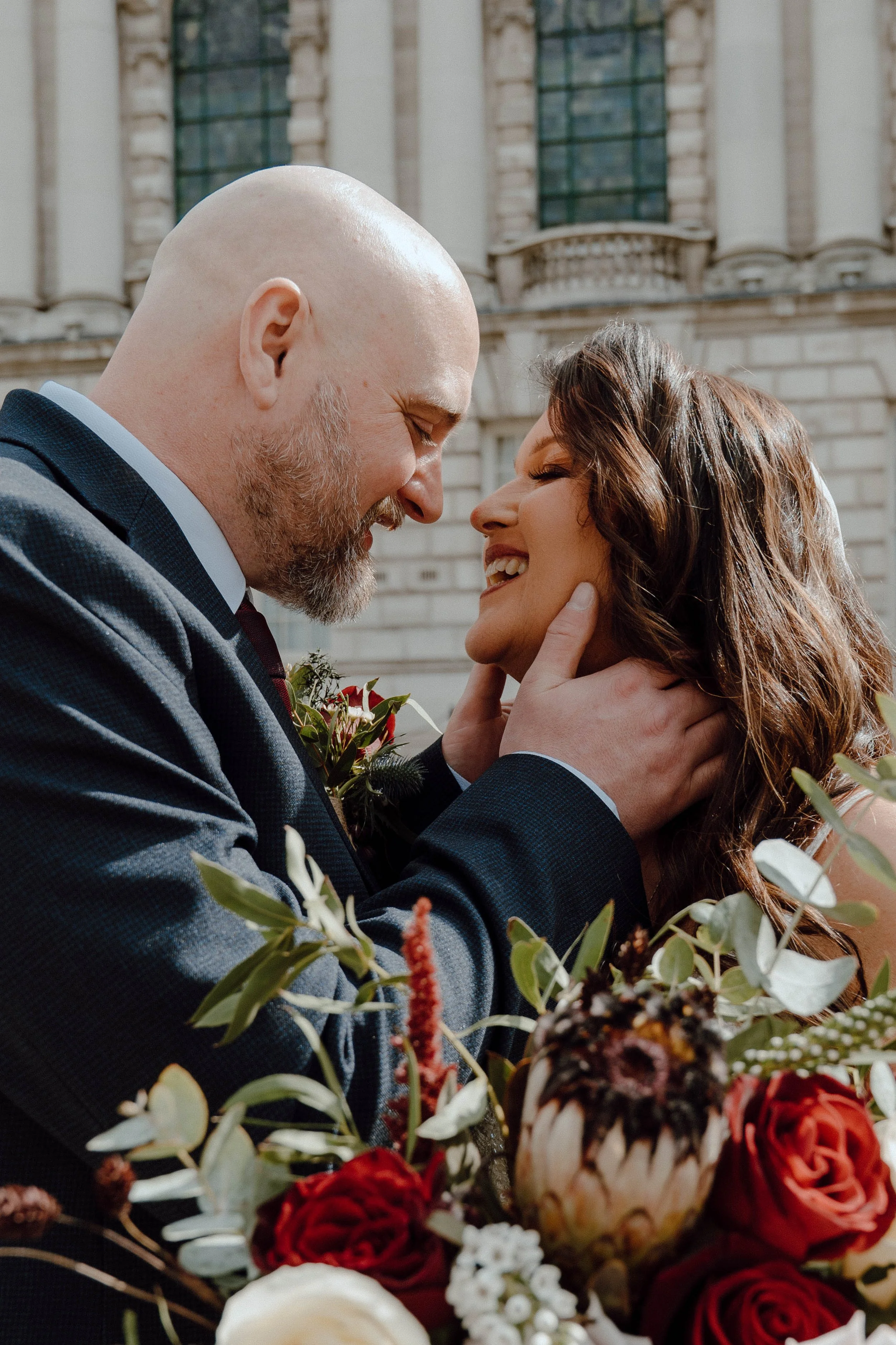 A happy bride and groom in wedding attire sharing an intimate moment with their foreheads touching and smiling, surrounded by a bouquet of flowers outdoors in front of a historic stone building.