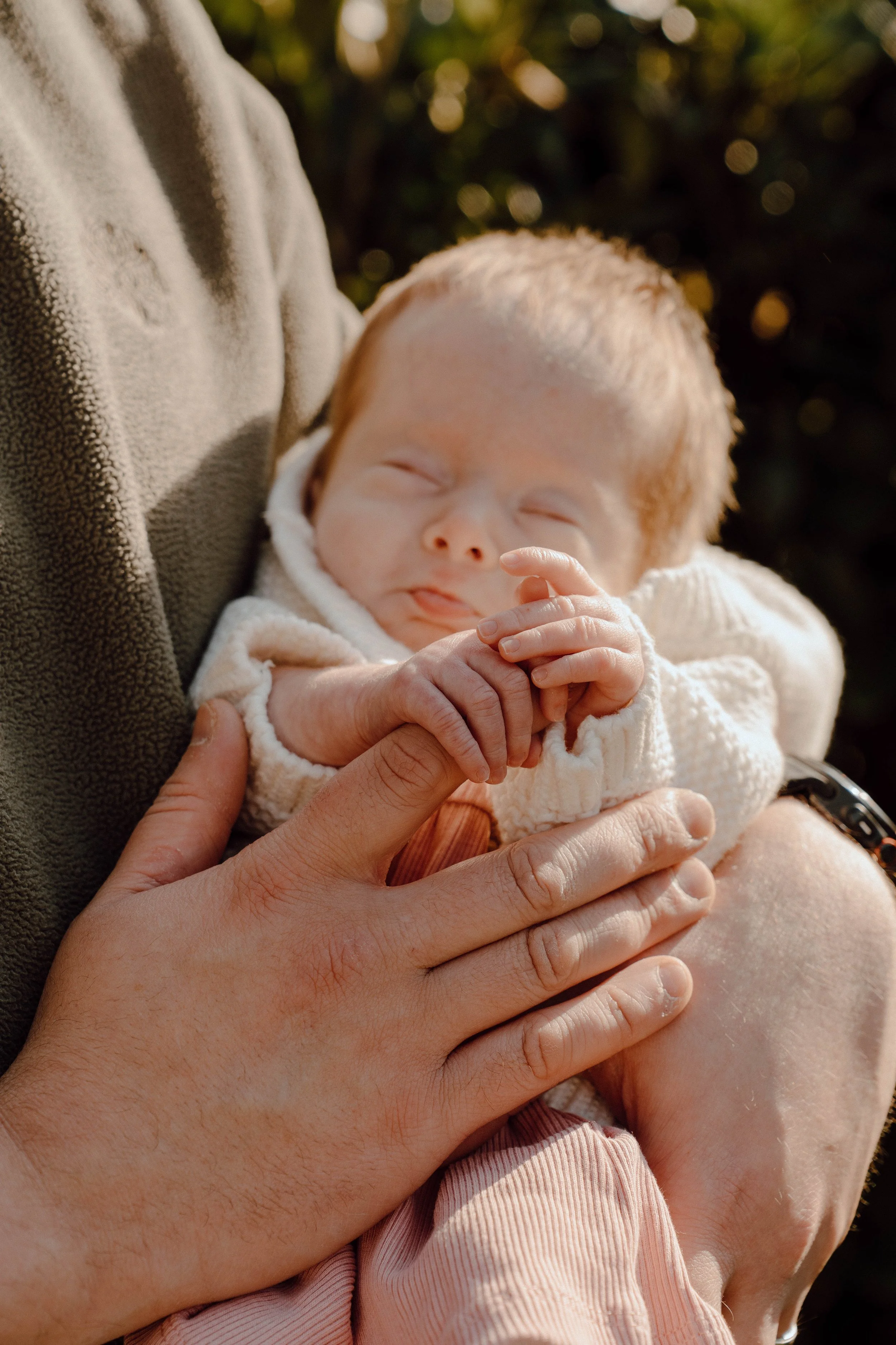 A sleeping baby with red hair and closed eyes is being held by an adult wearing a brown sweater, with a hand gently touching the baby's chest. The background is blurred with bokeh lights.