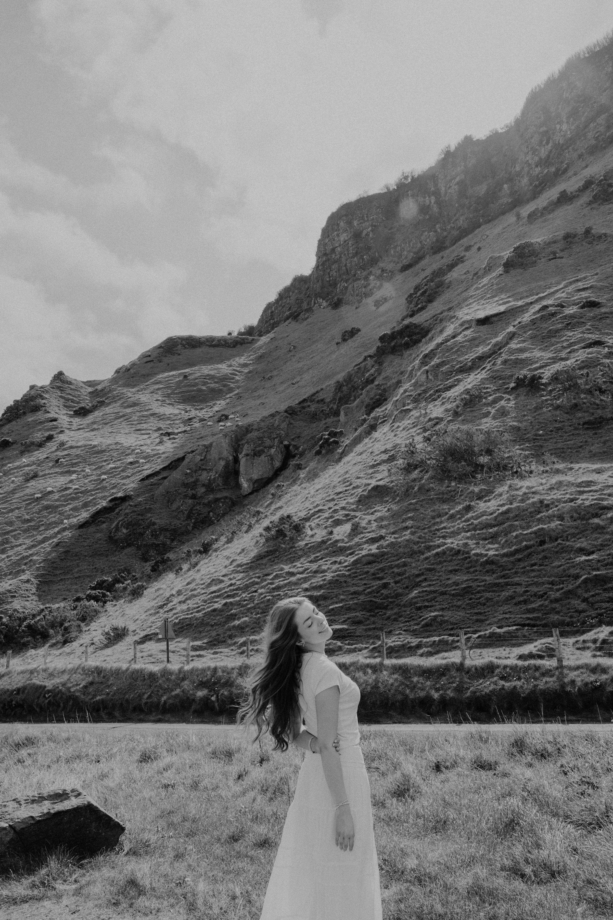 A woman in a white dress standing outdoors on grass with mountains in the background.