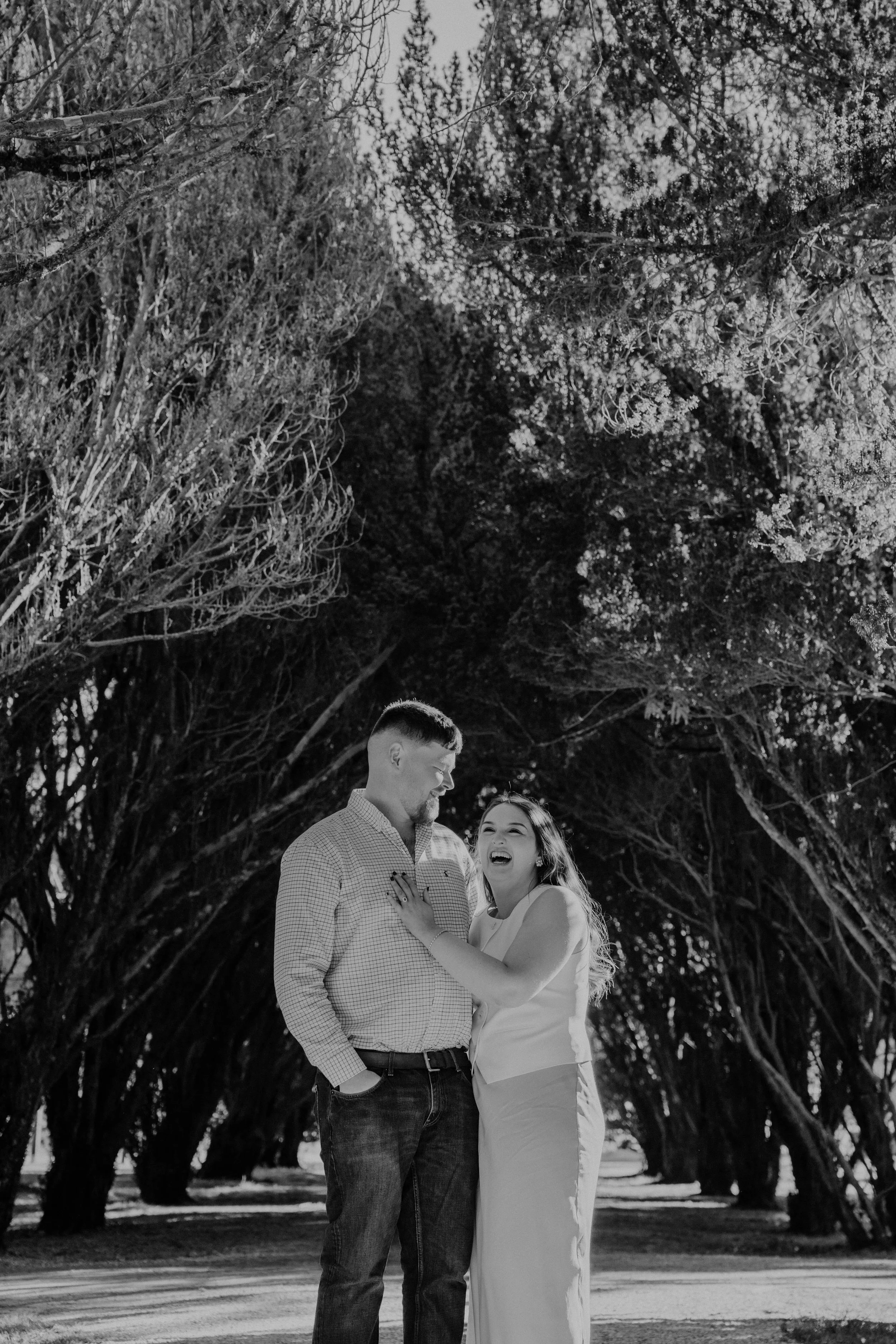 A smiling couple stands close together outdoors beneath a canopy of trees, sharing a joyful moment in black and white.