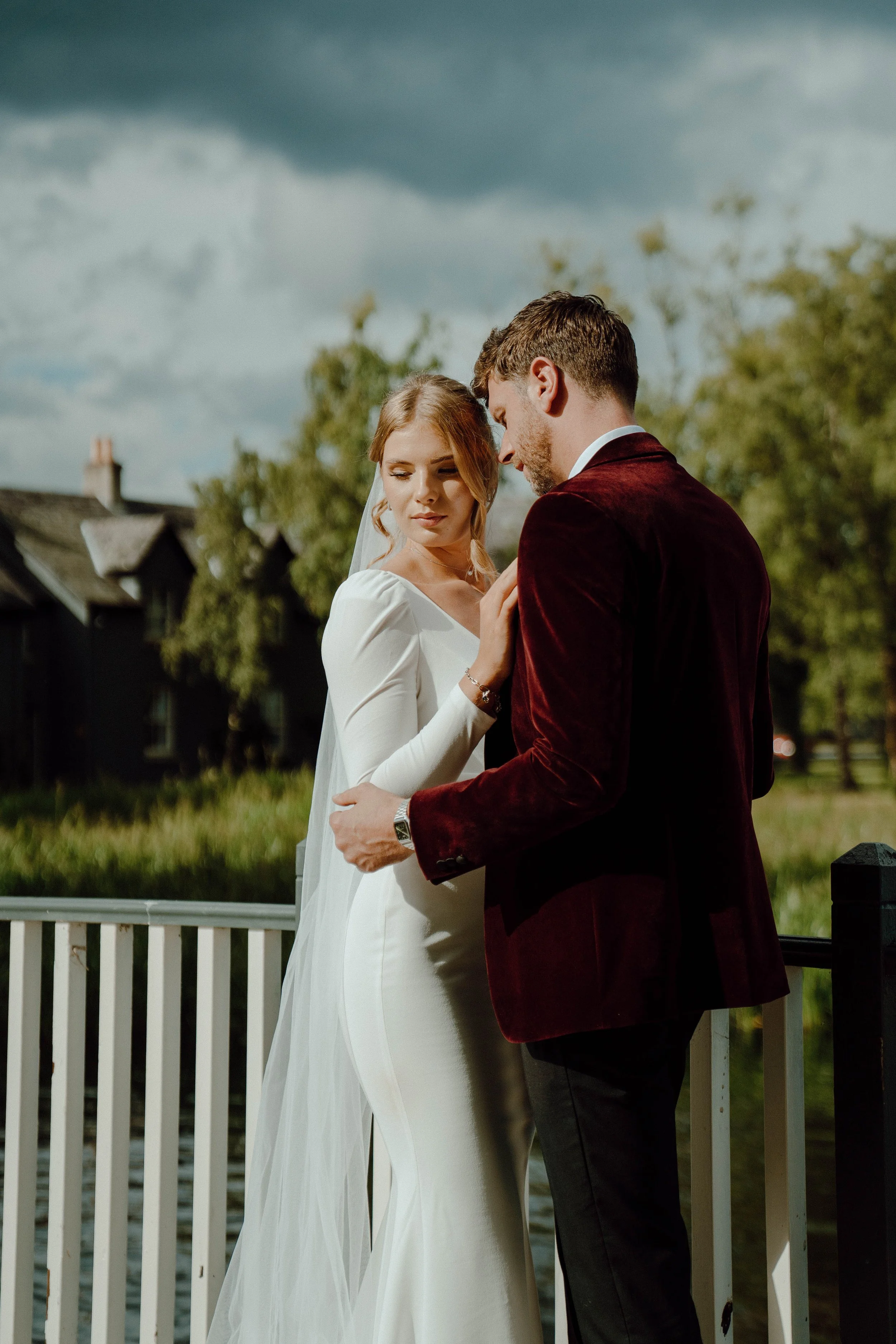A bride and groom stand close together outdoors, with a cloudy sky and trees in the background. The bride is in a white wedding dress with a veil, and the groom is in a dark red velvet blazer and dark pants.