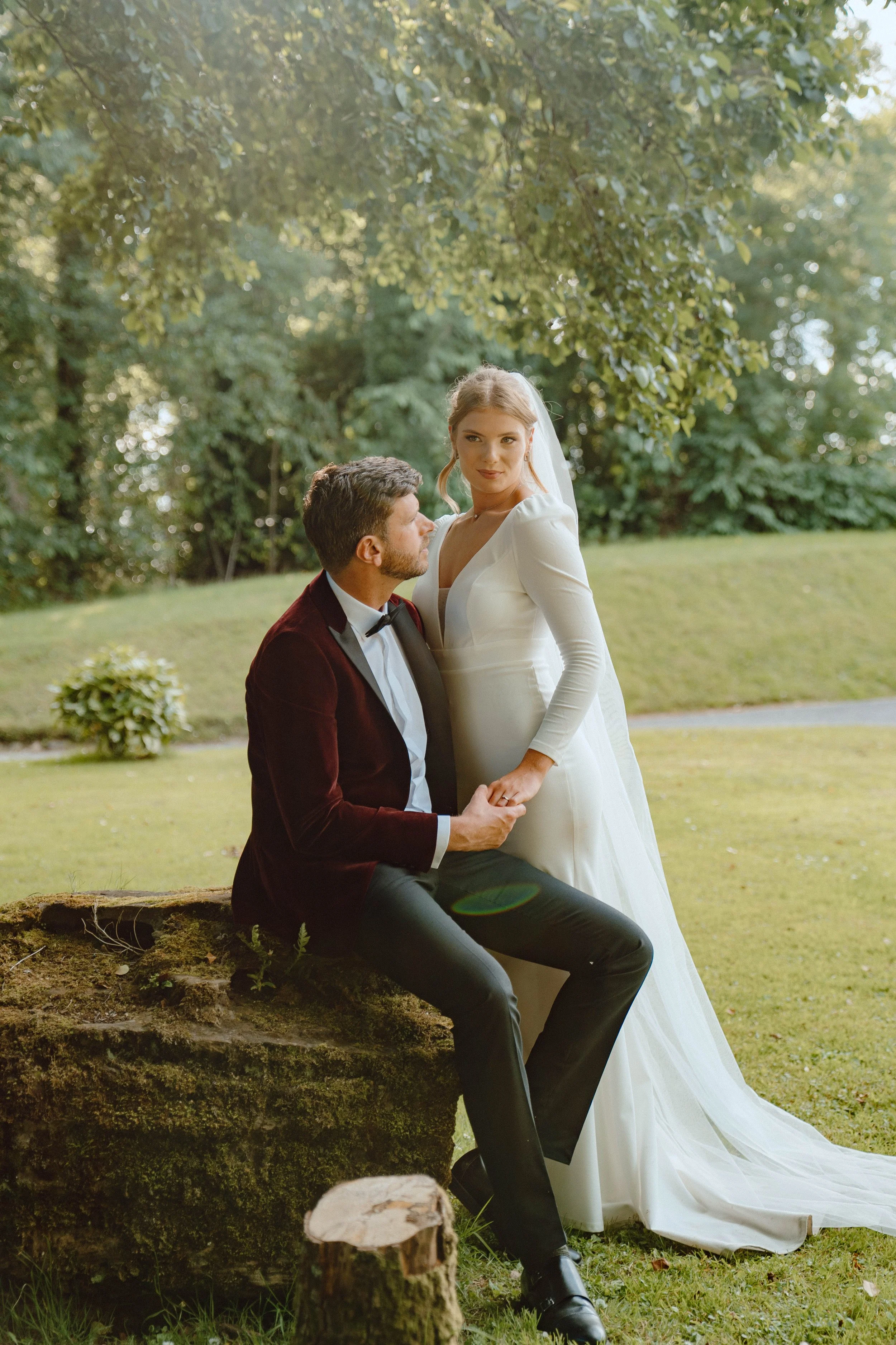 A bride and groom outdoors, with the groom seated on a tree stump and the bride standing beside him, holding his hand, surrounded by greenery and trees, during daytime.