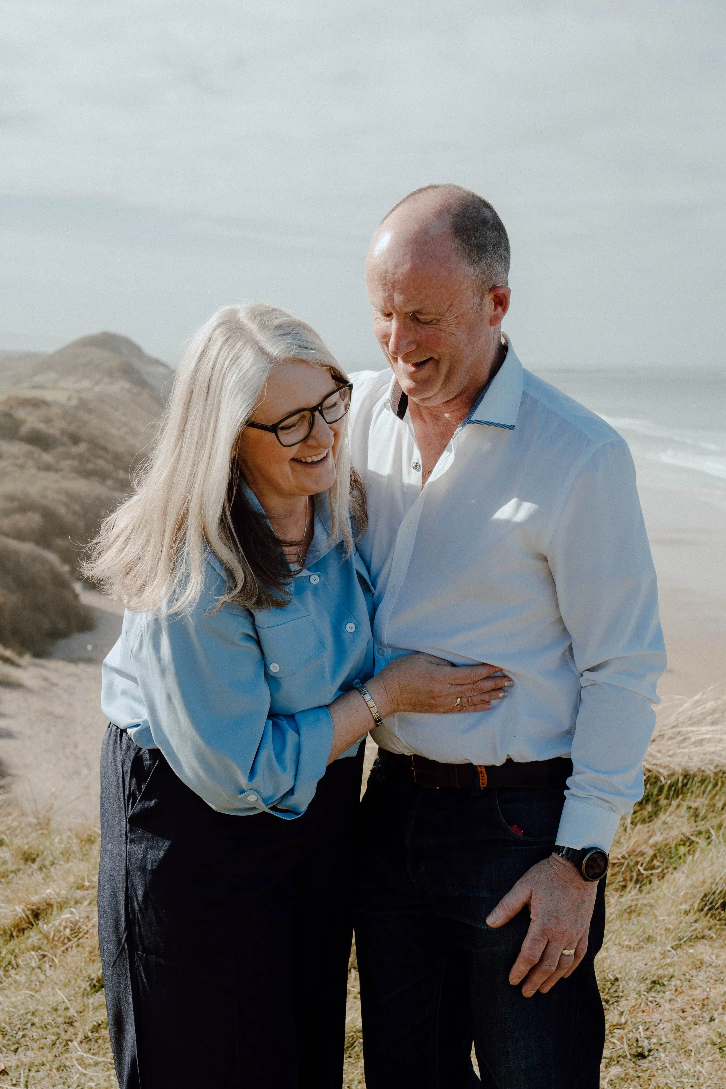 A smiling woman with glasses and long gray hair wearing a light blue shirt, embracing a smiling man with a buzz cut and wearing a white shirt, on a beach with dunes and ocean in the background.