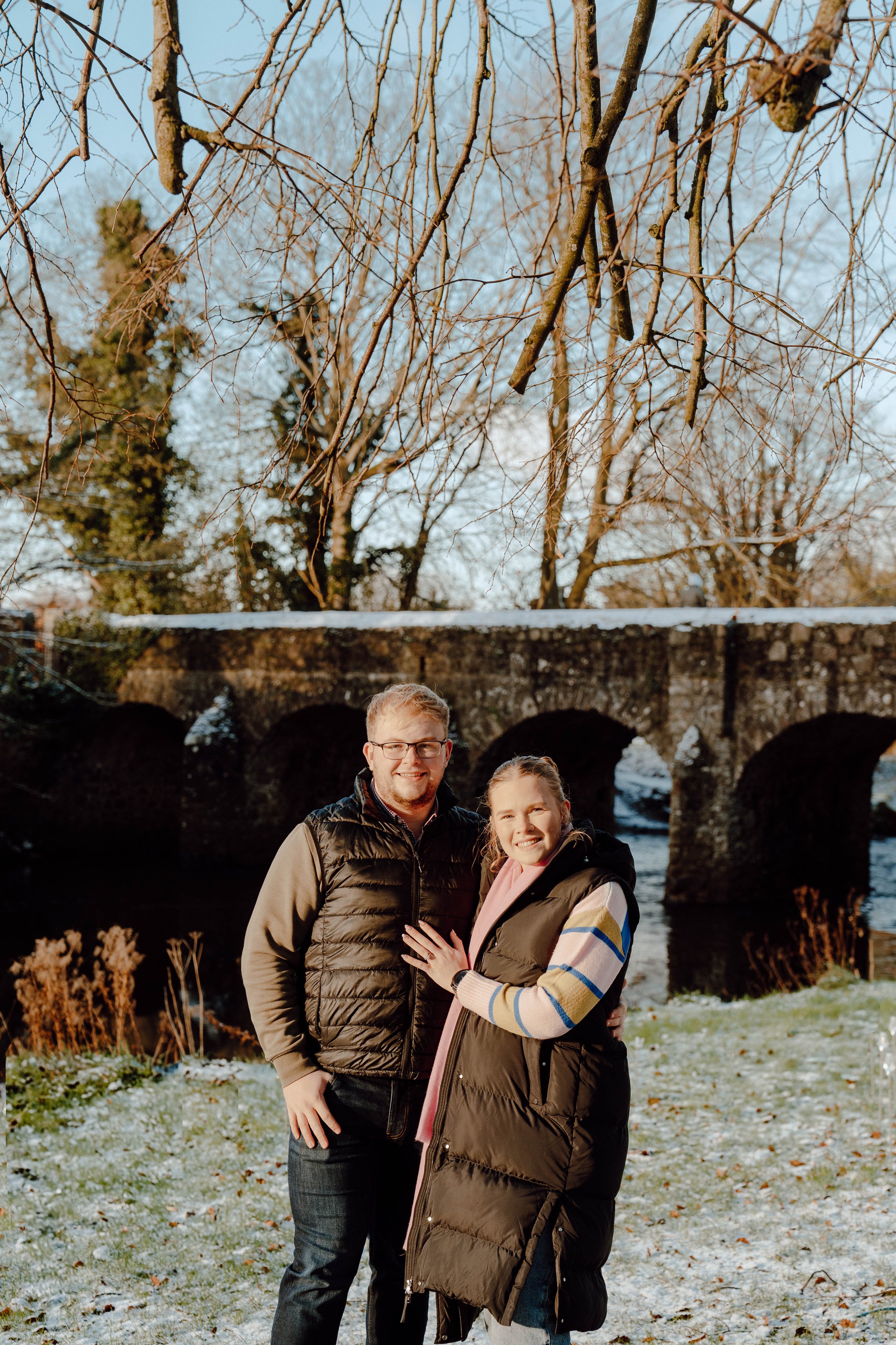 A man and woman smiling outdoors in front of a stone bridge with water flowing underneath, on a winter day with some snow and leafless trees.