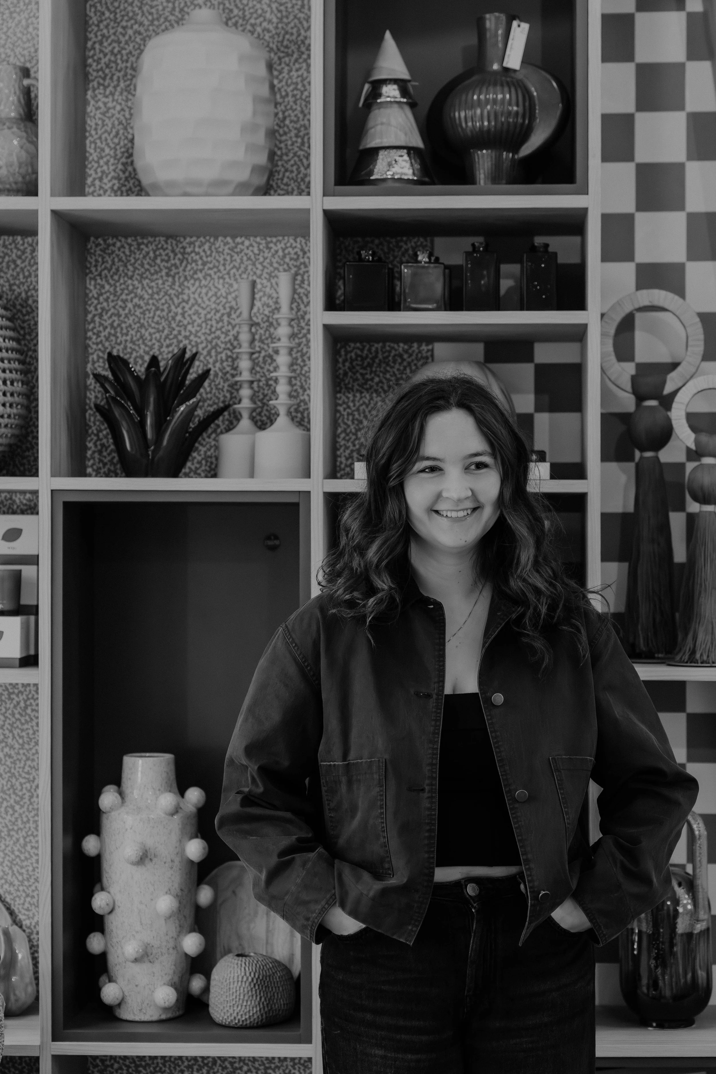 A young woman with wavy hair, smiling, wearing a dark jacket and black top, standing in front of a bookshelf filled with decorative objects.
