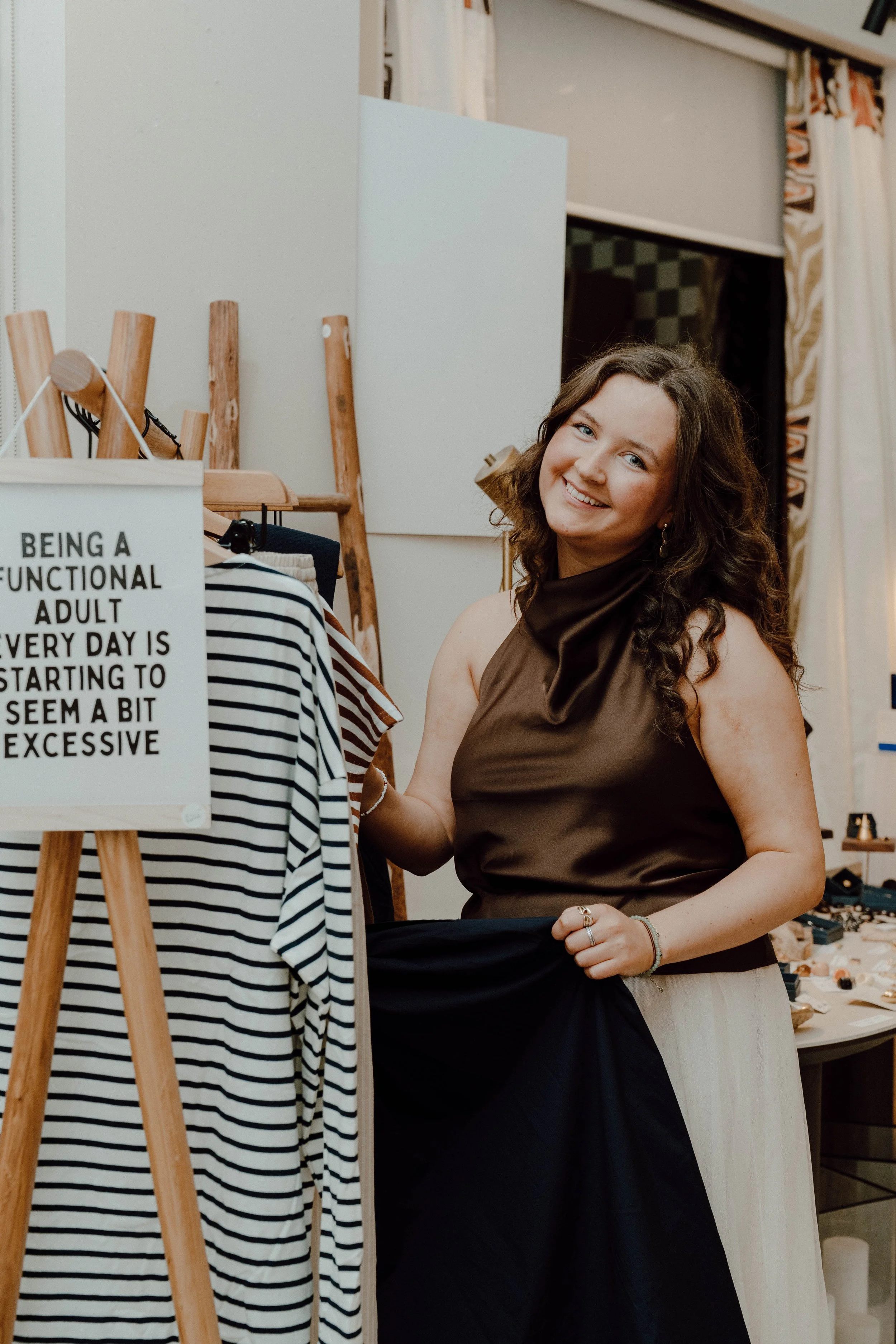A woman with wavy brown hair wearing a brown sleeveless top and a beige skirt, smiling and shopping for striped clothing in a boutique store.