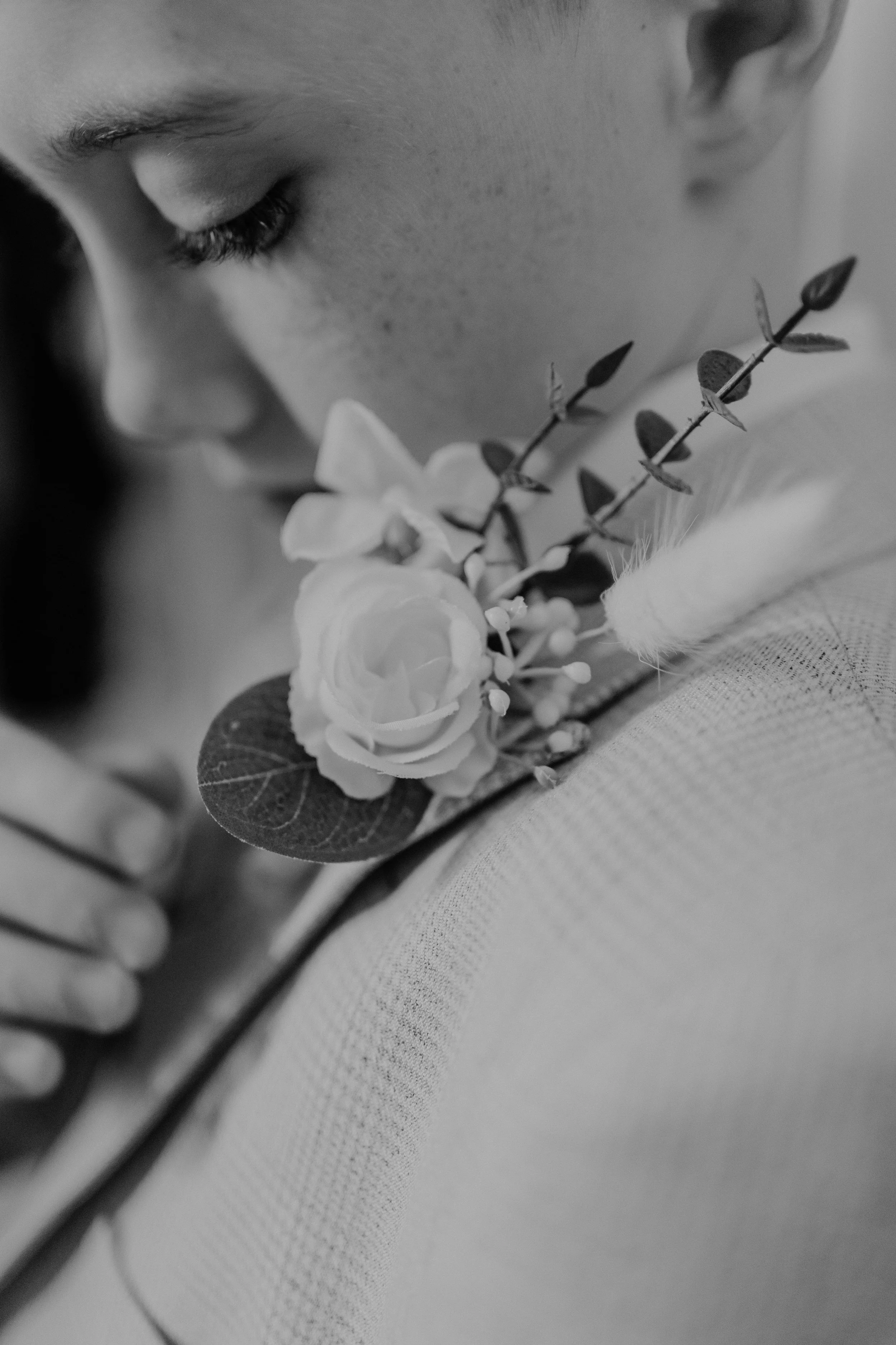Close-up of a person's face near a boutonniere with white rose, eucalyptus leaves, and other small flowers, on a textured fabric surface in black and white.