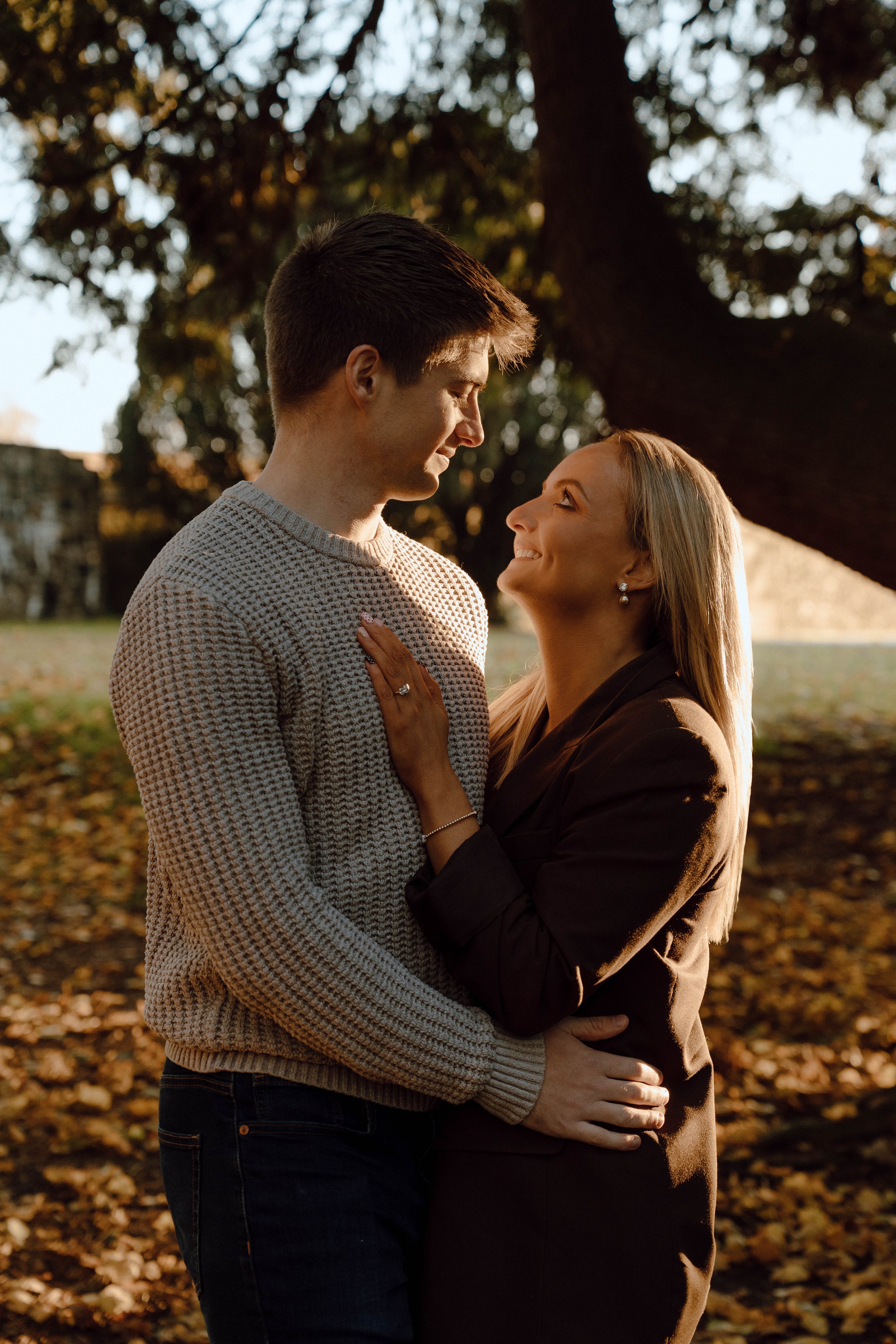 A couple standing close together outdoors under a large tree during autumn, smiling and looking into each other's eyes, with fallen leaves on the ground.