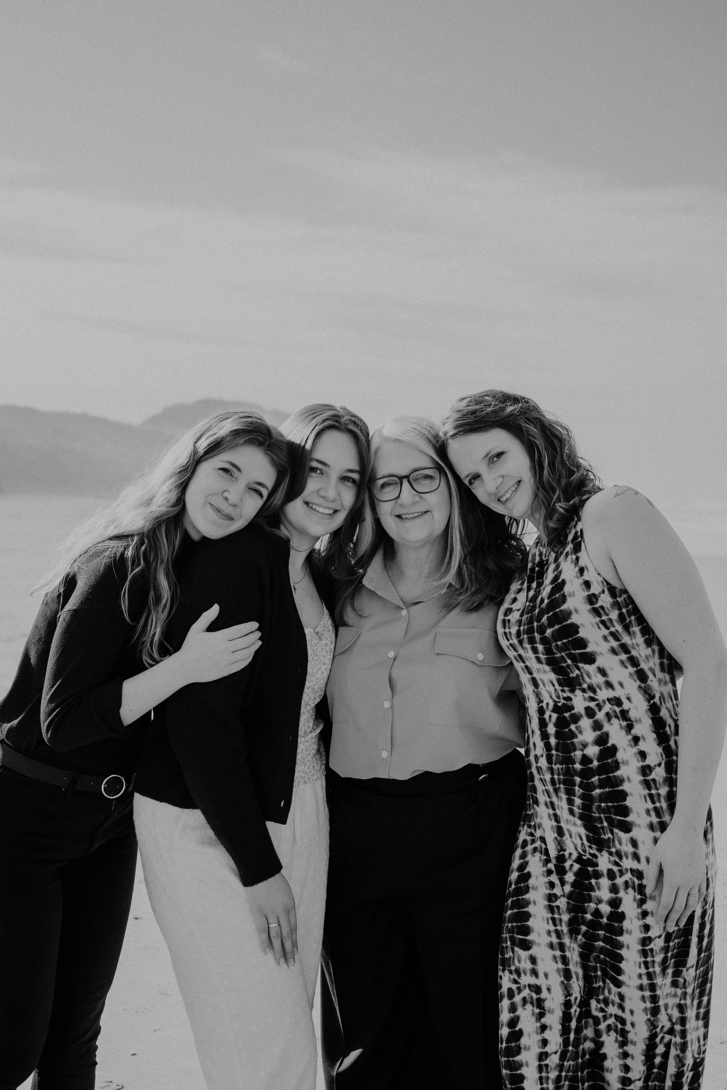 A group of four women standing close together outdoors, smiling, with a scenic backdrop of mountains and sky in black and white.