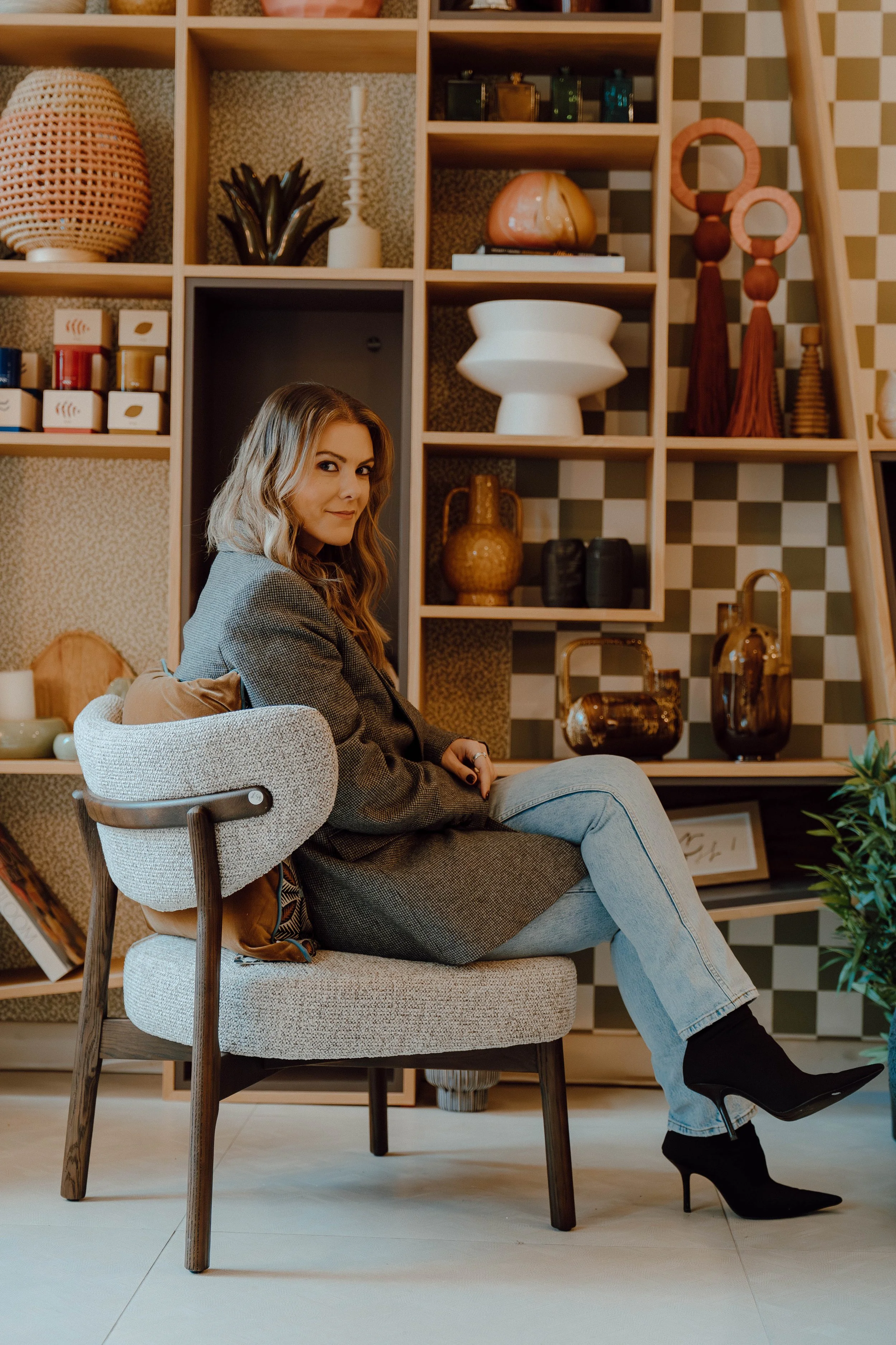 A woman with light brown hair sitting on a modern chair in front of a decorative wooden shelf filled with vases, sculptures, and books.