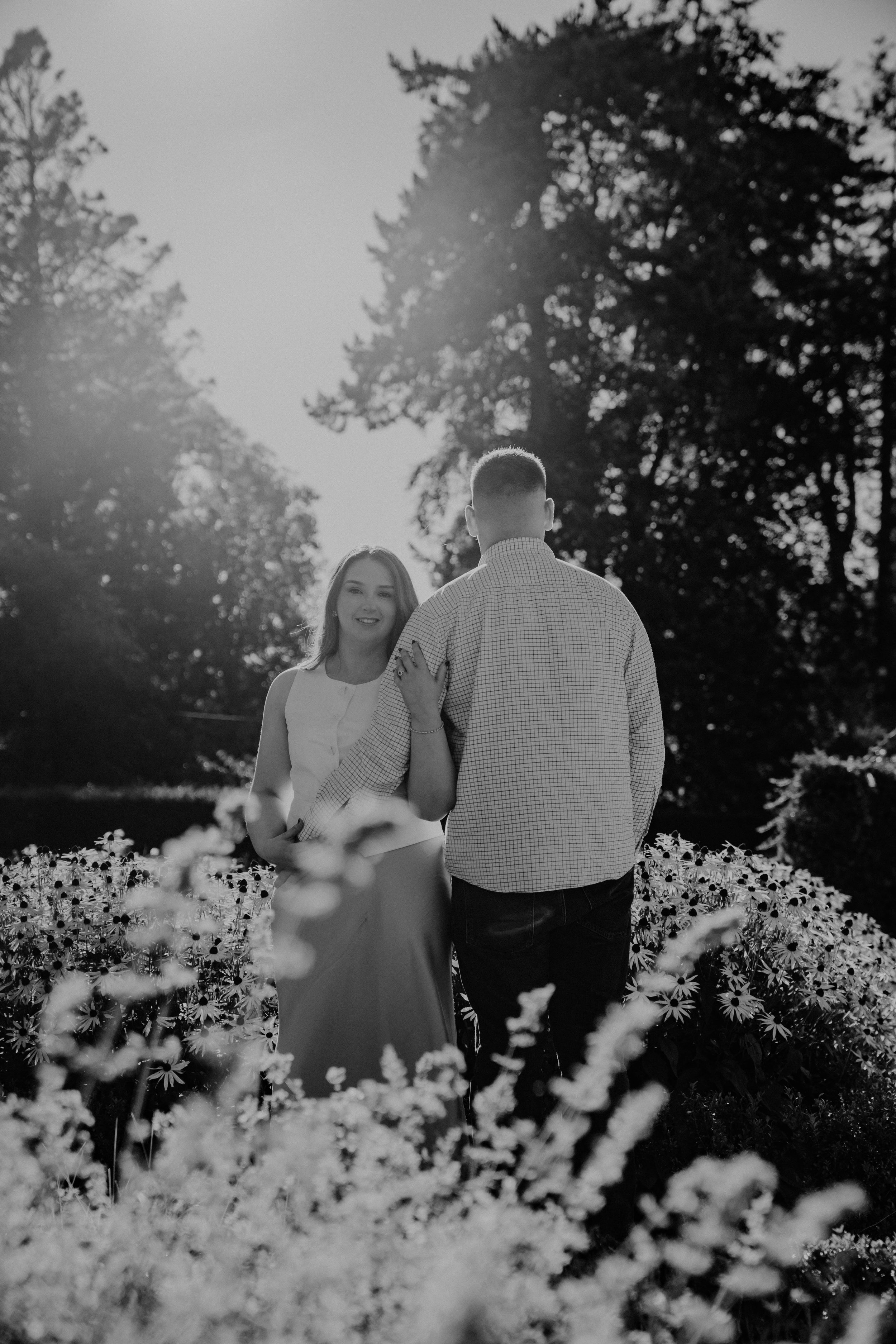 A woman and a man stand in a garden full of flowers, with sunlight shining through trees behind them. The woman is smiling and holding the man's arm.