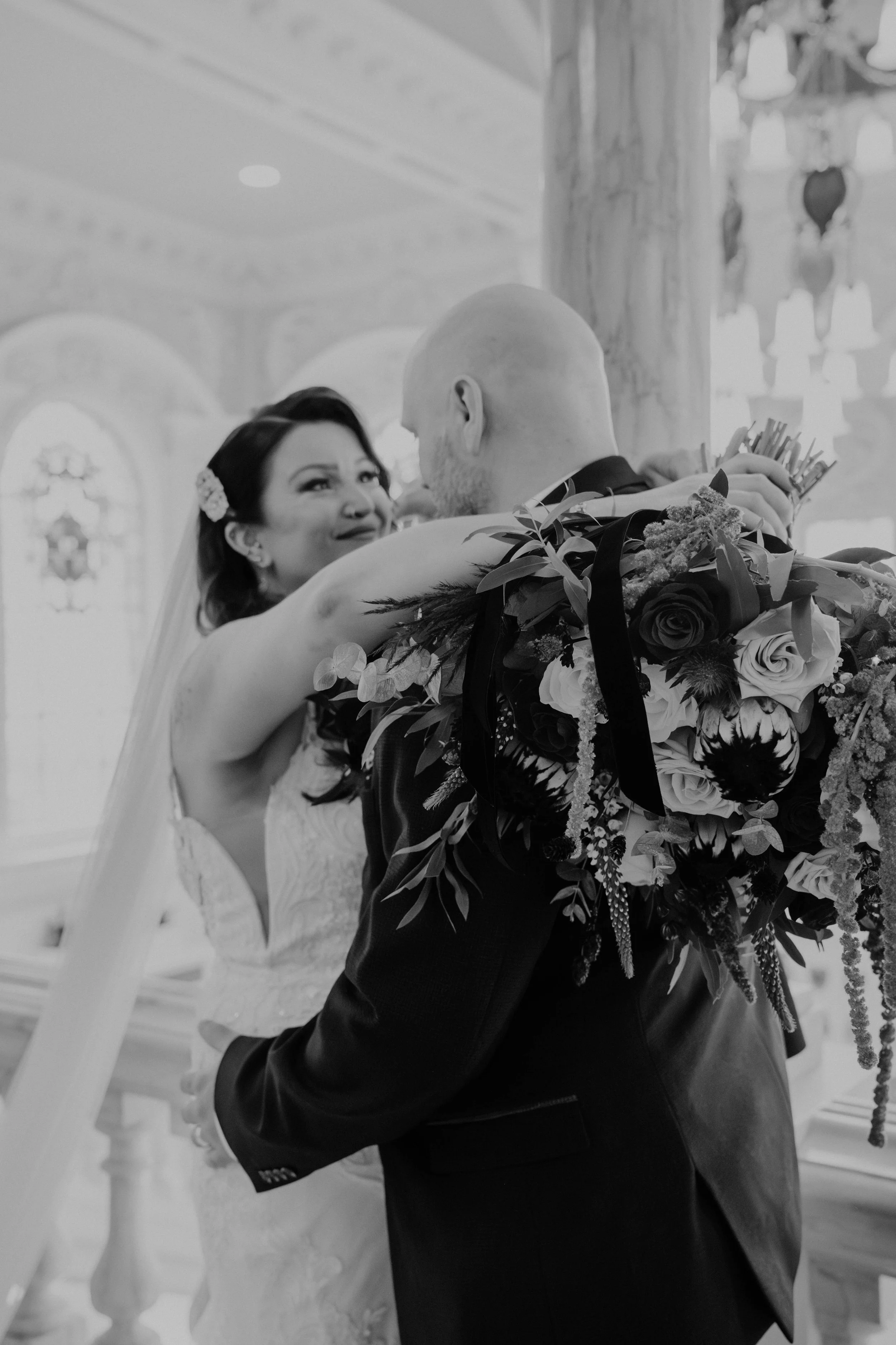 Black and white photo of a bride and groom embracing during a wedding, with the bride smiling and holding a large bouquet of flowers.