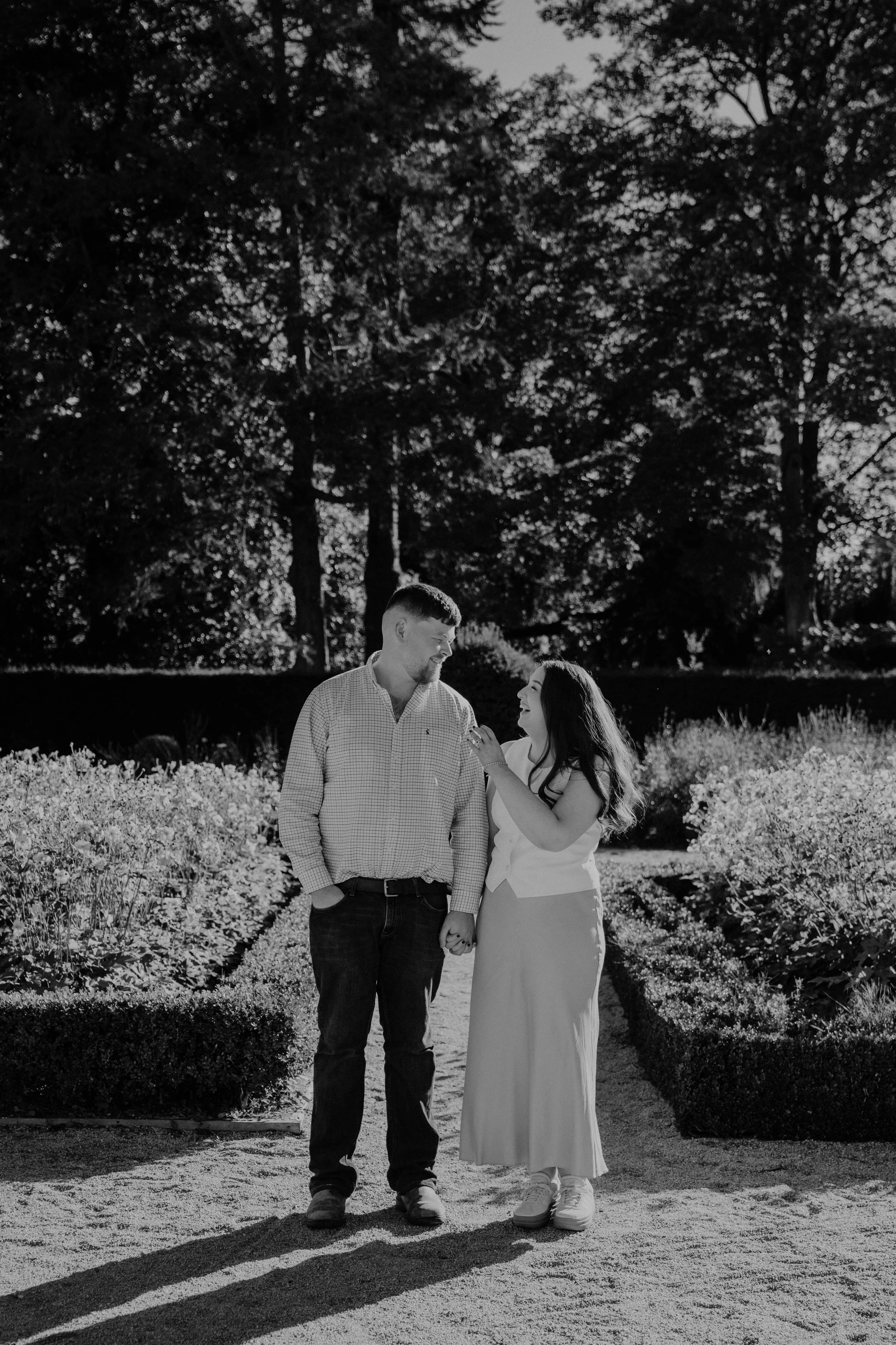 A black and white photo of a couple holding hands, walking in a flower garden with tall trees in the background, smiling at each other.