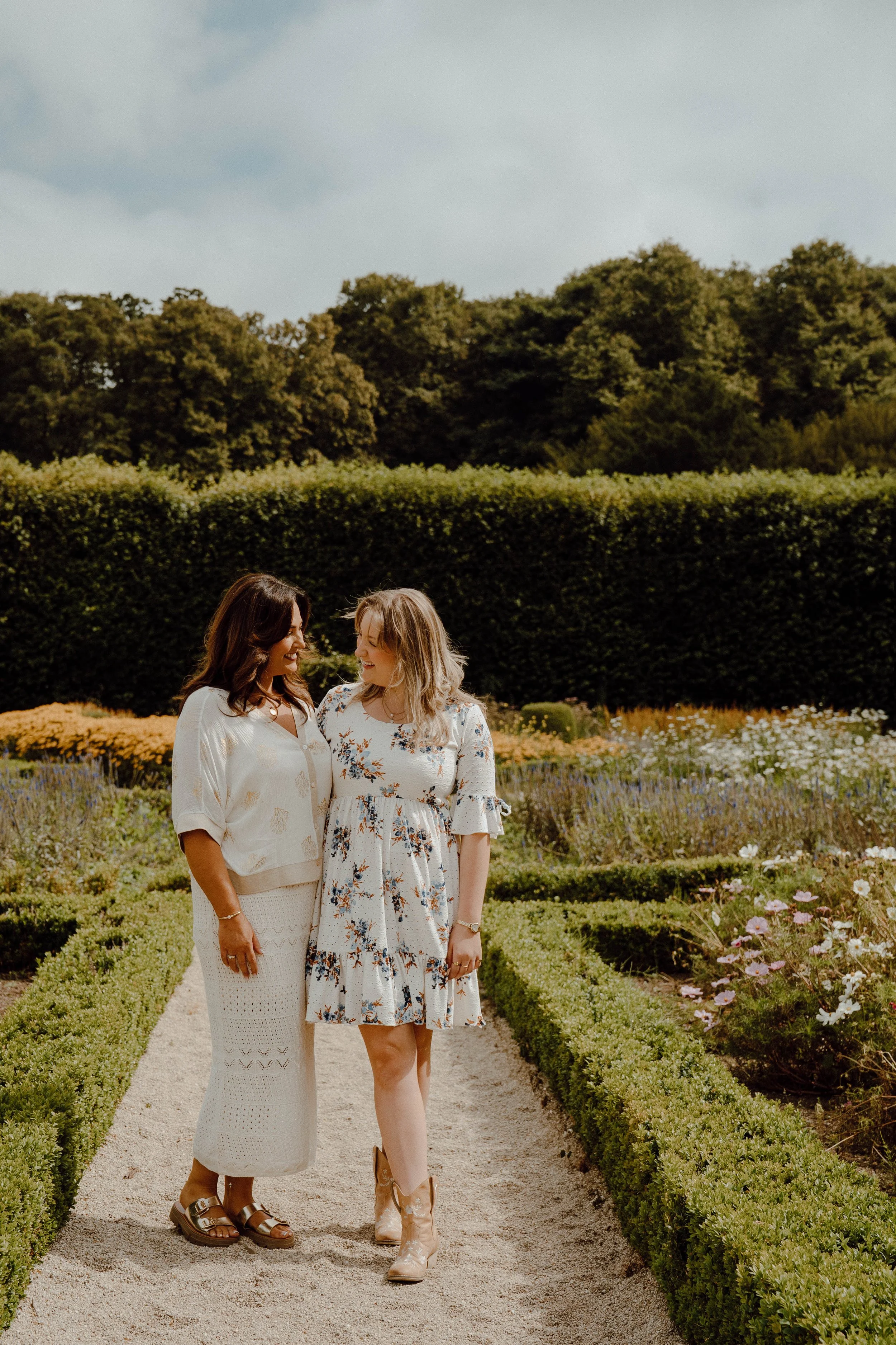 Two women walking and talking in a garden with flowering plants and green shrubbery, under a cloudy sky.