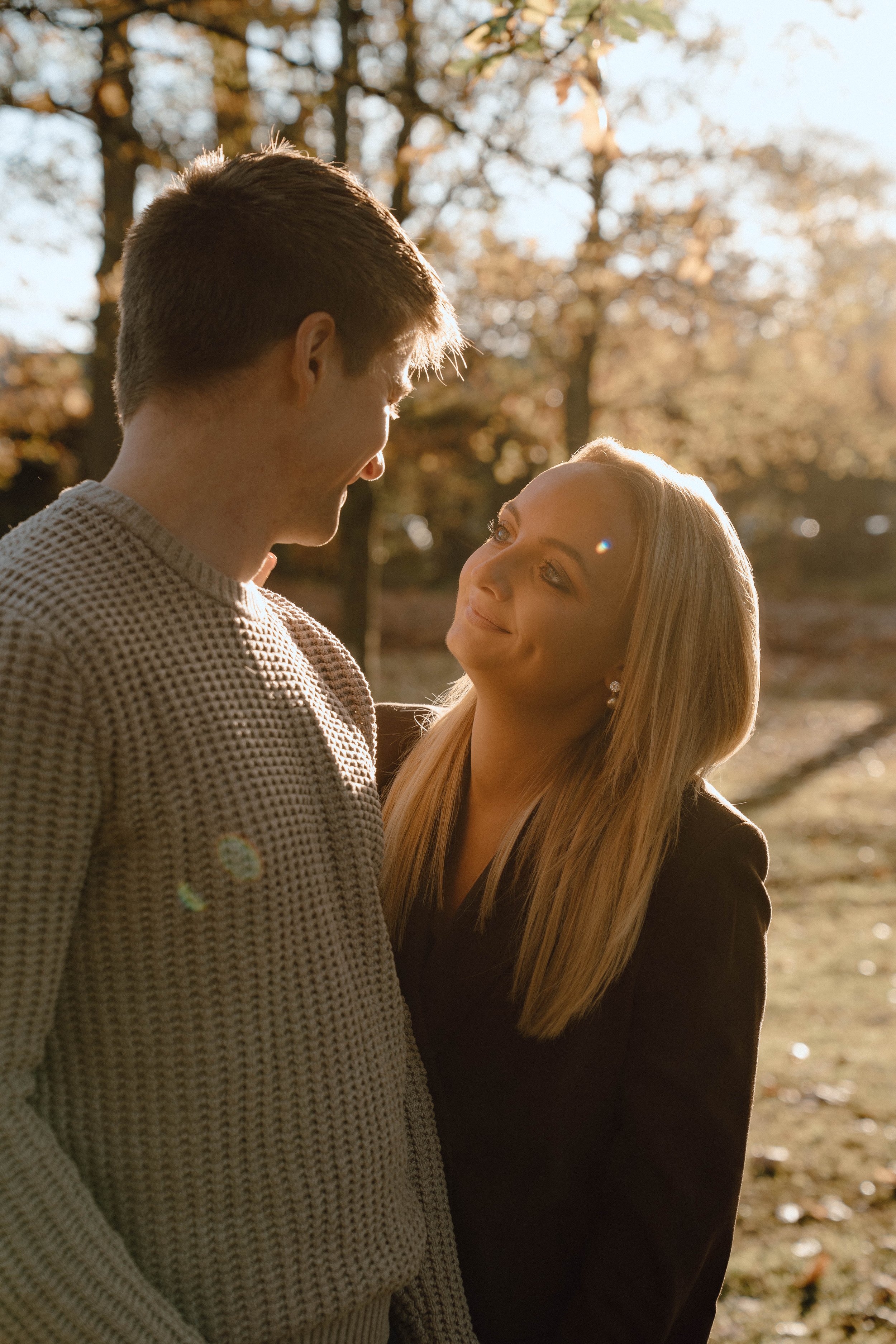 A man and woman looking at each other outdoors in sunlight with fall foliage in background.