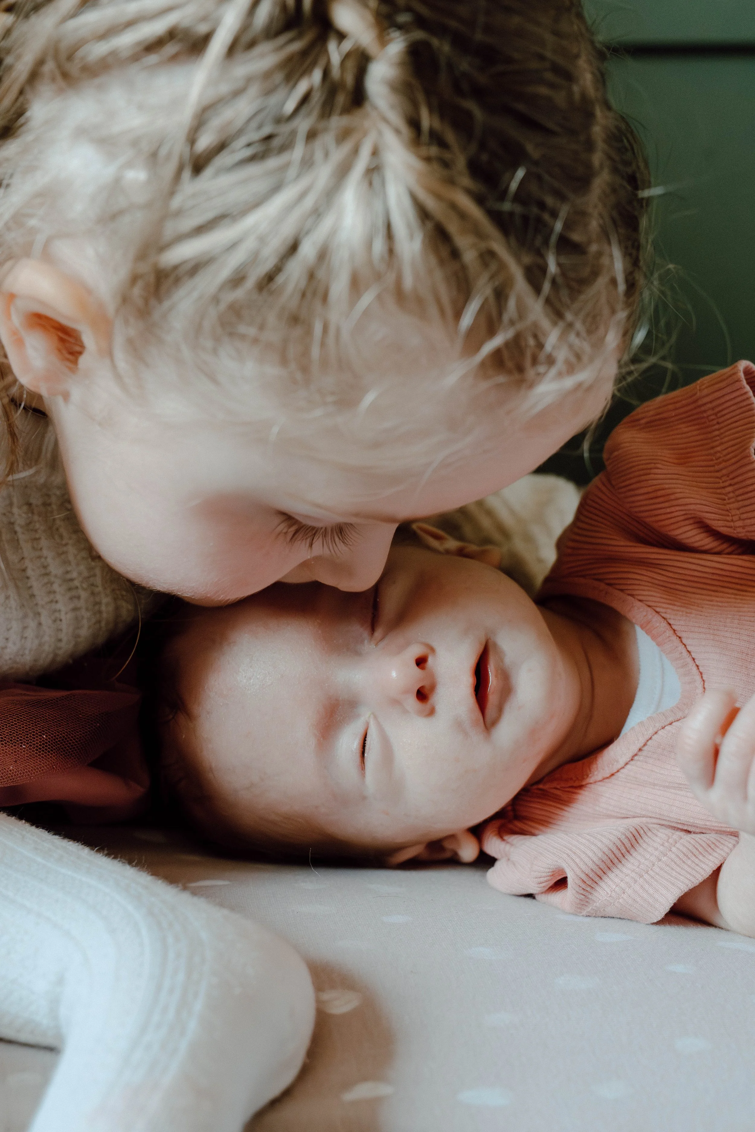 A young girl with wet hair leaning over a sleeping baby lying on its back on a white blanket.