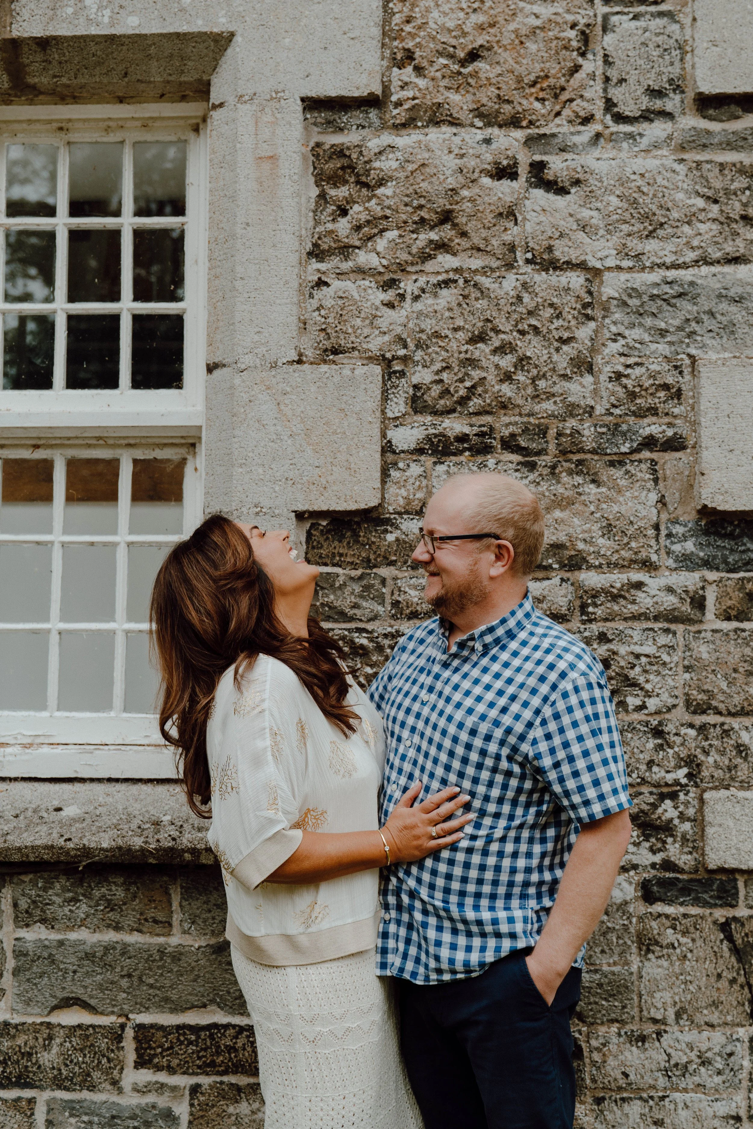 A smiling woman and man standing outdoors near a stone wall with a window, sharing a joyful moment.