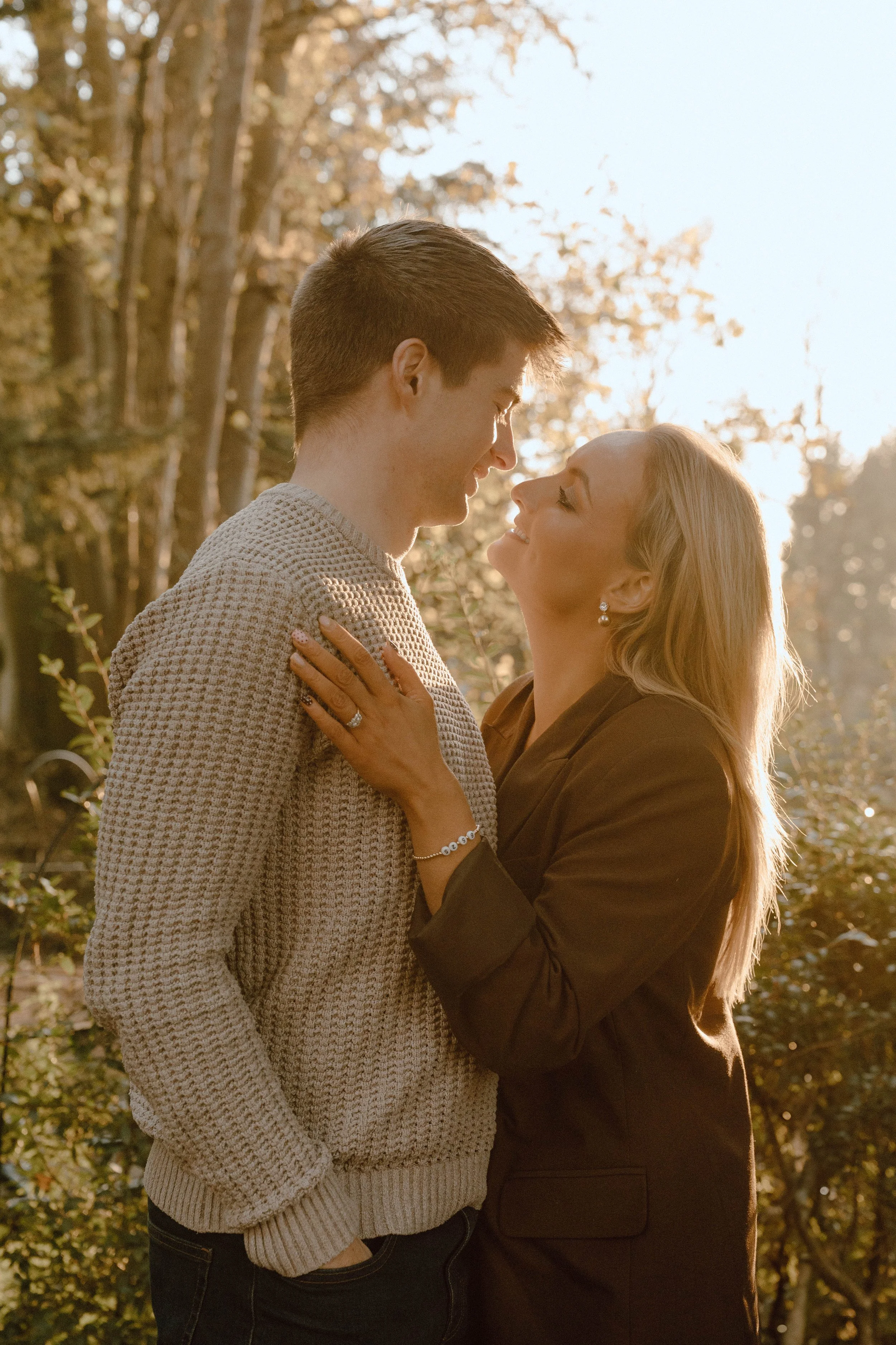 A young man and a woman are standing outdoors, gazing into each other's eyes, with trees and sunlight in the background.