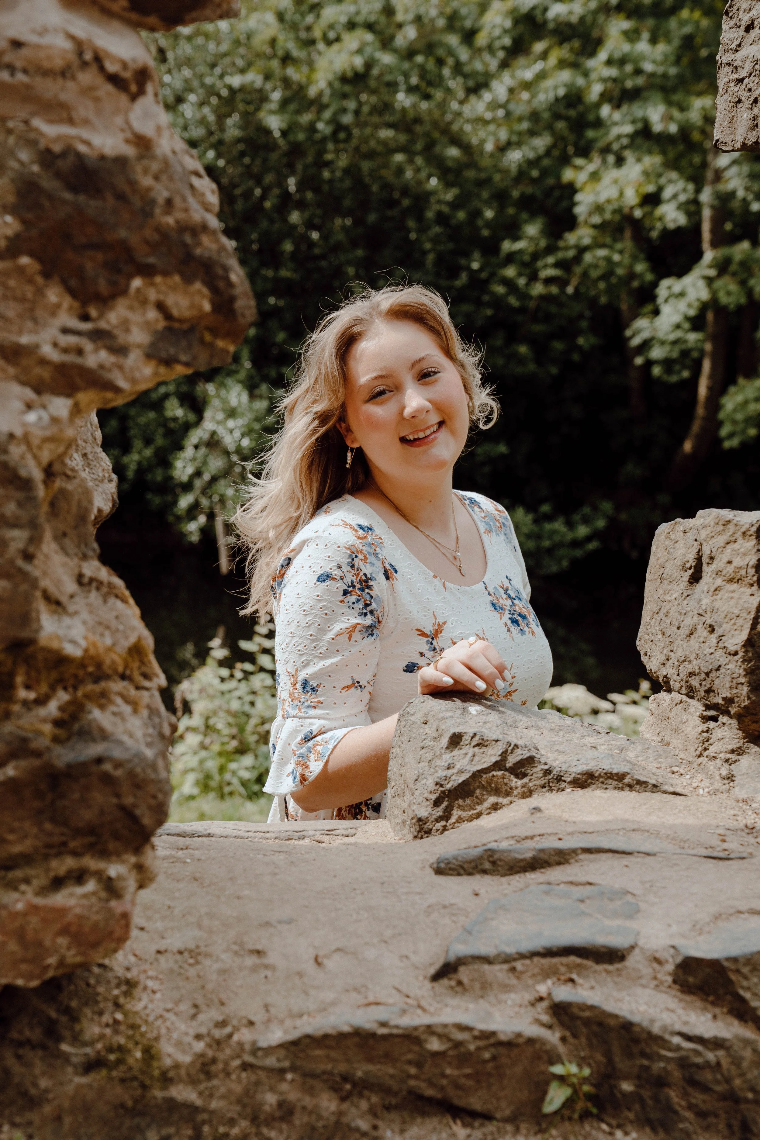 A young woman with wavy blonde hair standing outdoors between two stone walls, smiling at the camera, with greenery and trees in the background.