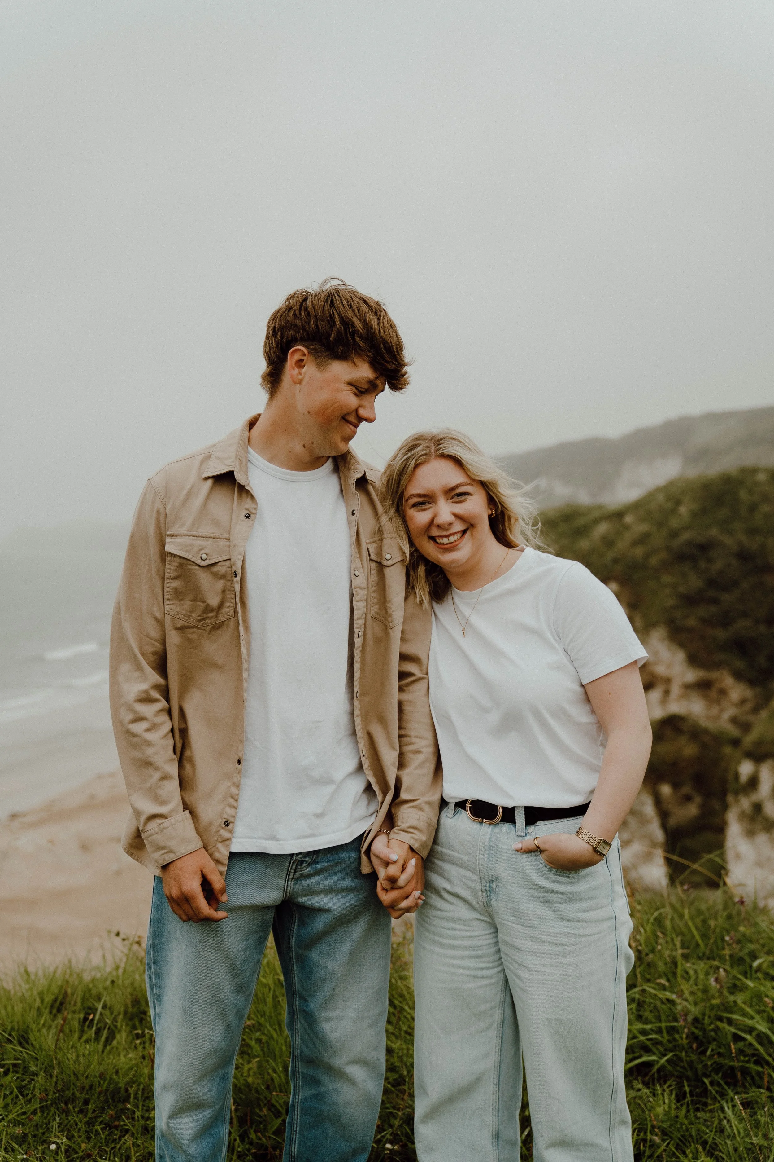 A smiling couple standing close together on a grassy area near a beach with cliffs in the background, holding hands and looking at each other.