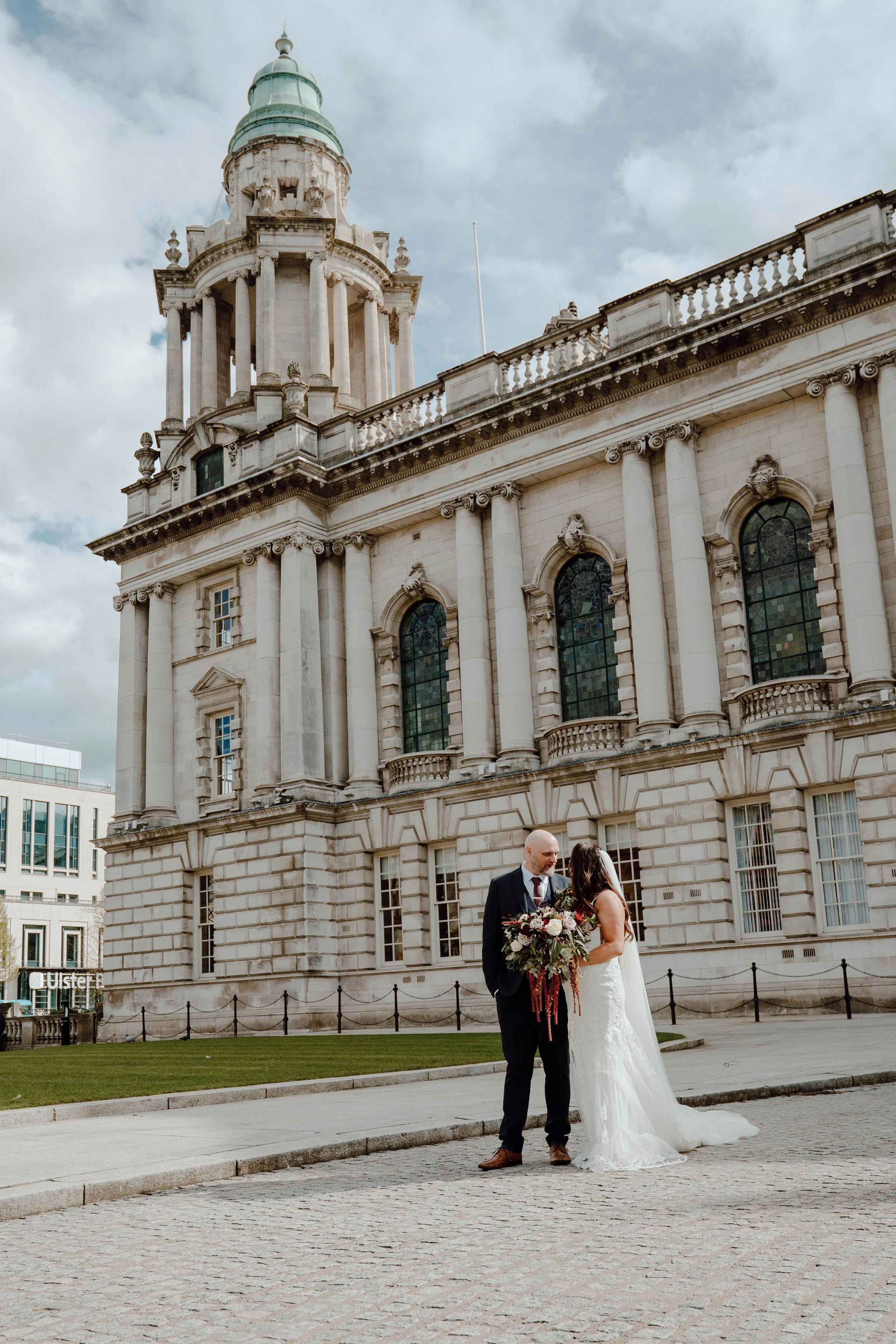 A bride and groom standing close together outside a historic building with large windows and ornate architectural details, on a cloudy day.