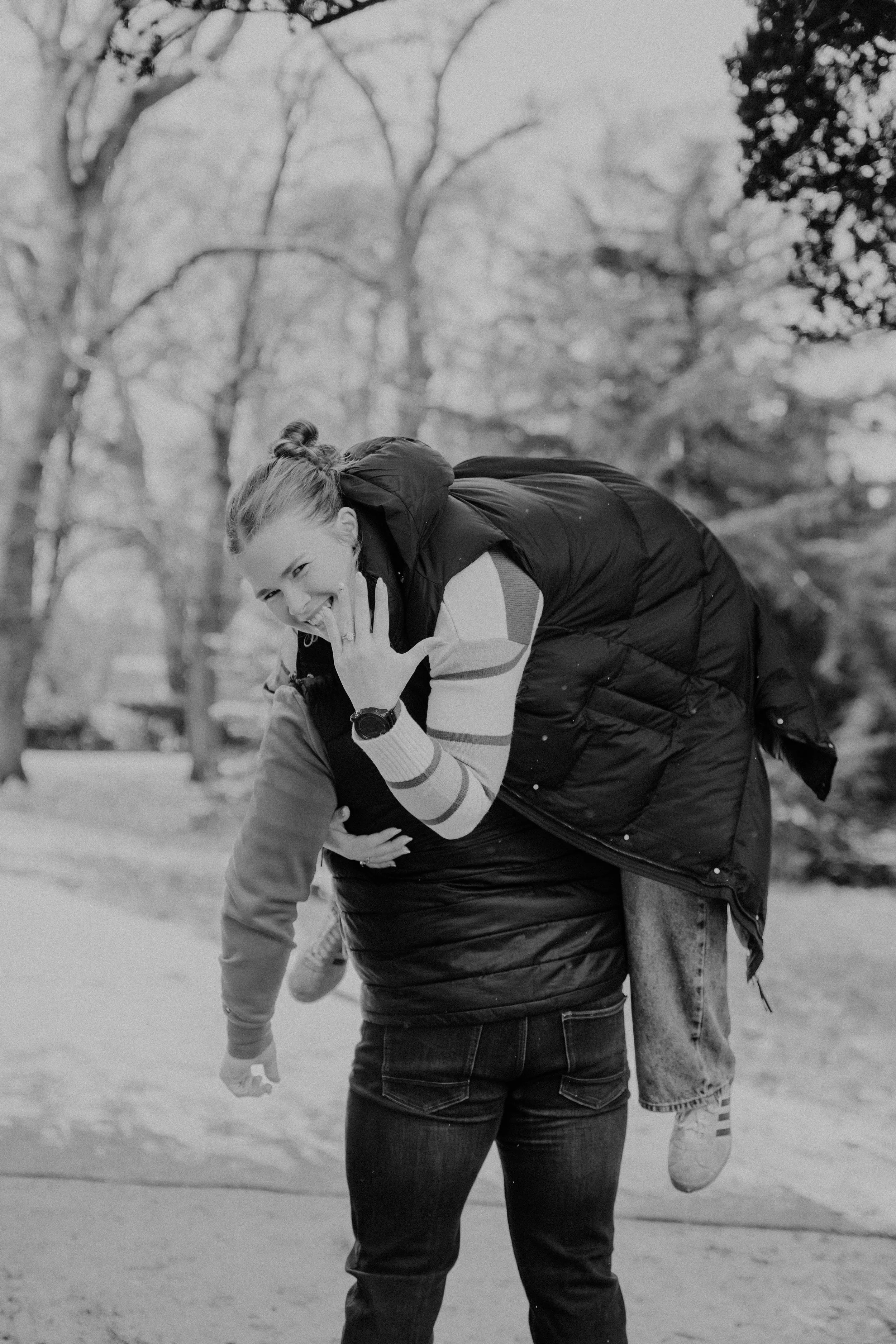 Woman playfully giving a piggyback ride outdoors, smiling and making a gesture with her hand, during daytime in a park with trees in the background, in black and white.