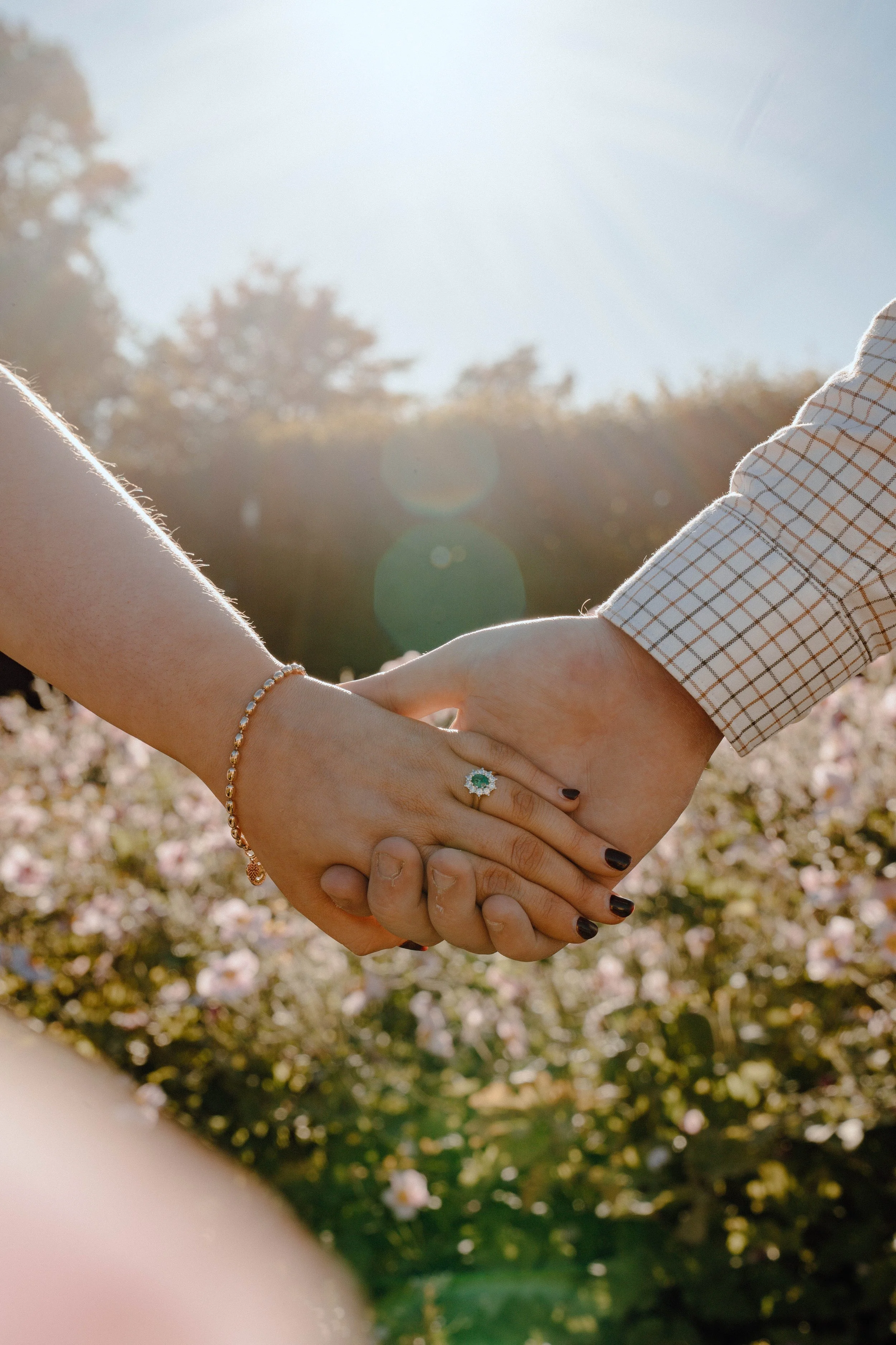 Close-up of a couple holding hands outdoors during daytime, with sunlight shining, in front of a backdrop of flowering bushes and trees.