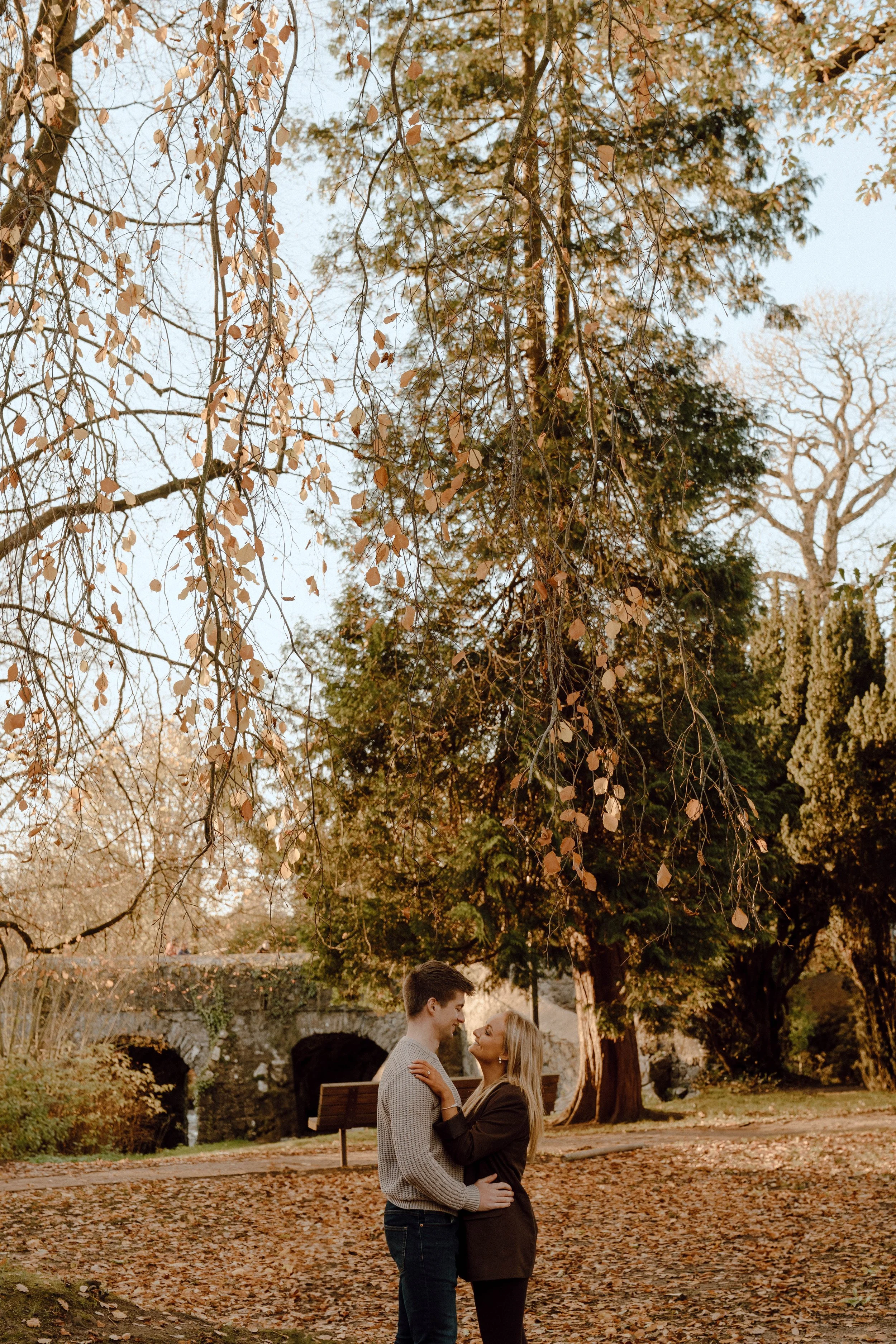 A couple in love standing in a park during autumn, embracing each other, with fallen leaves on the ground and trees in the background.