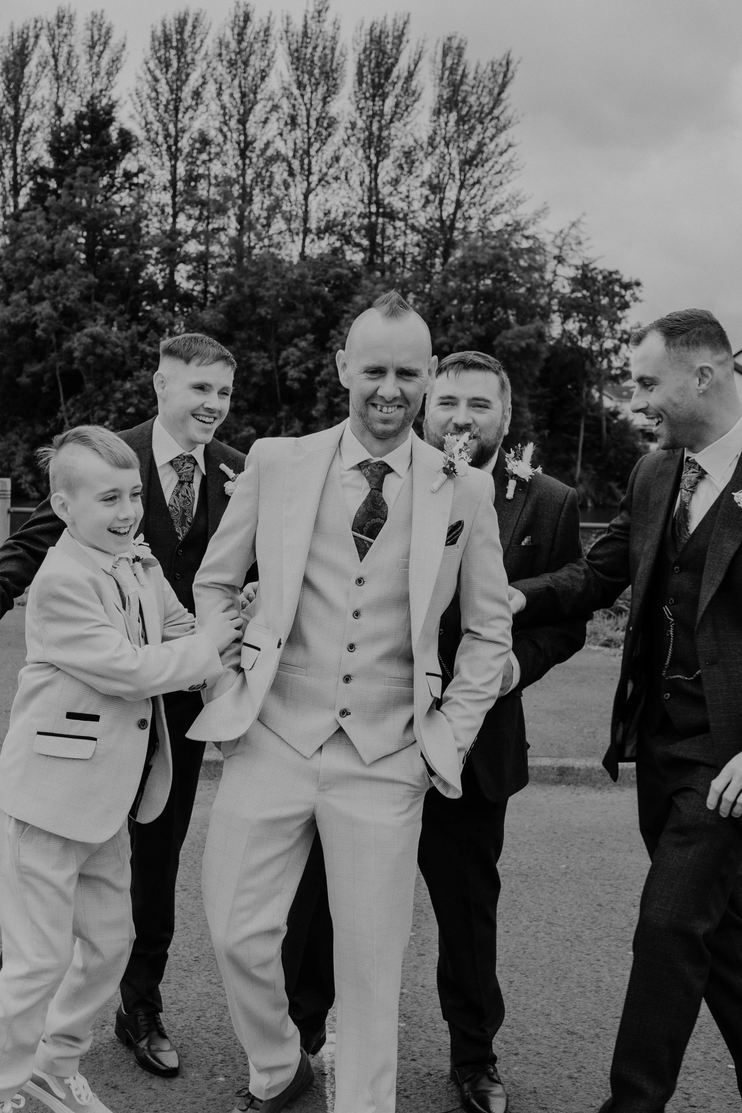 A black and white photo of a groom surrounded by five men and boys, all dressed in suits. They are outdoors, smiling and happy.