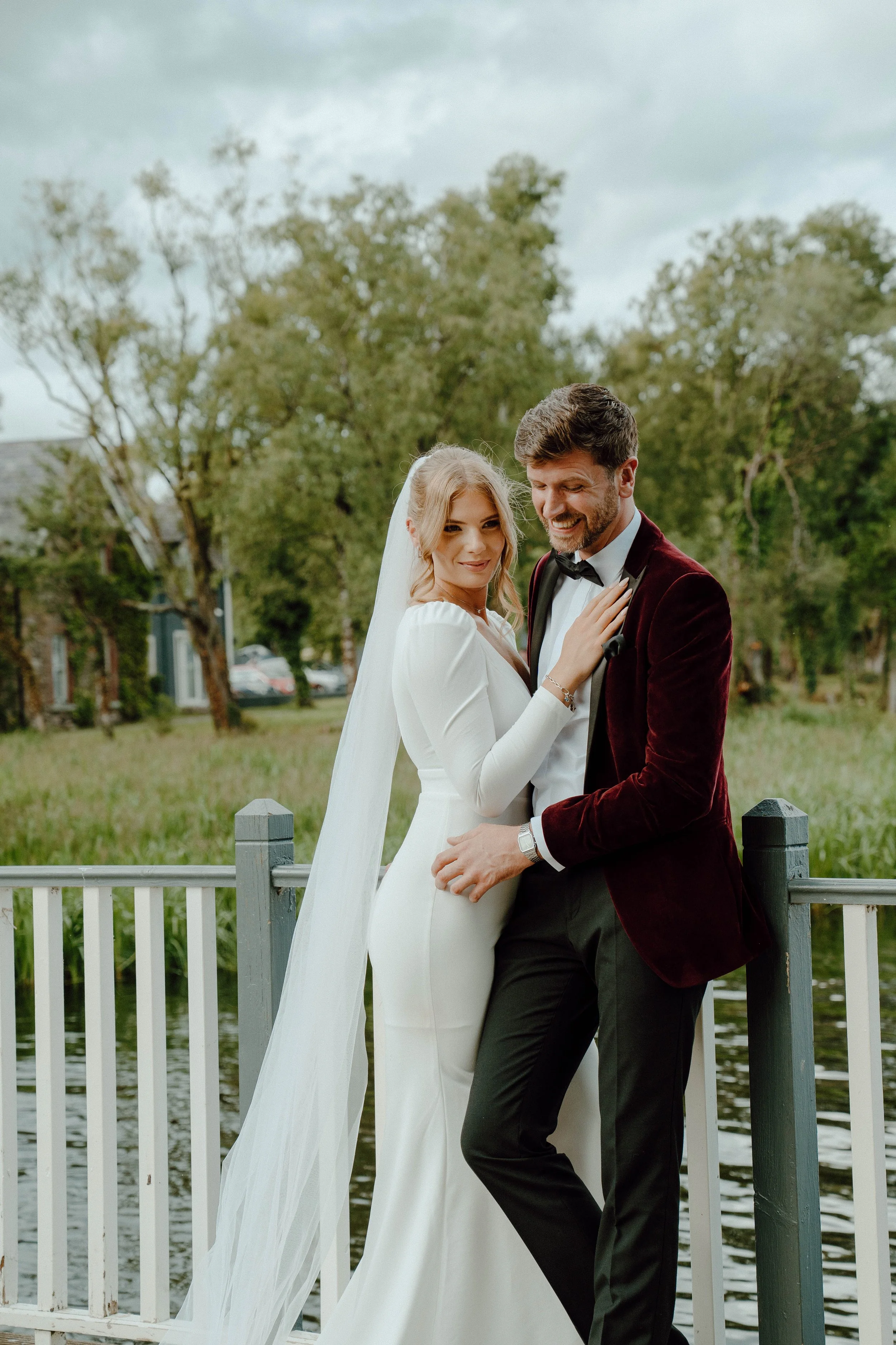 Bride and groom standing on a bridge outdoors, with trees and a grassy field in the background, during a wedding photoshoot.