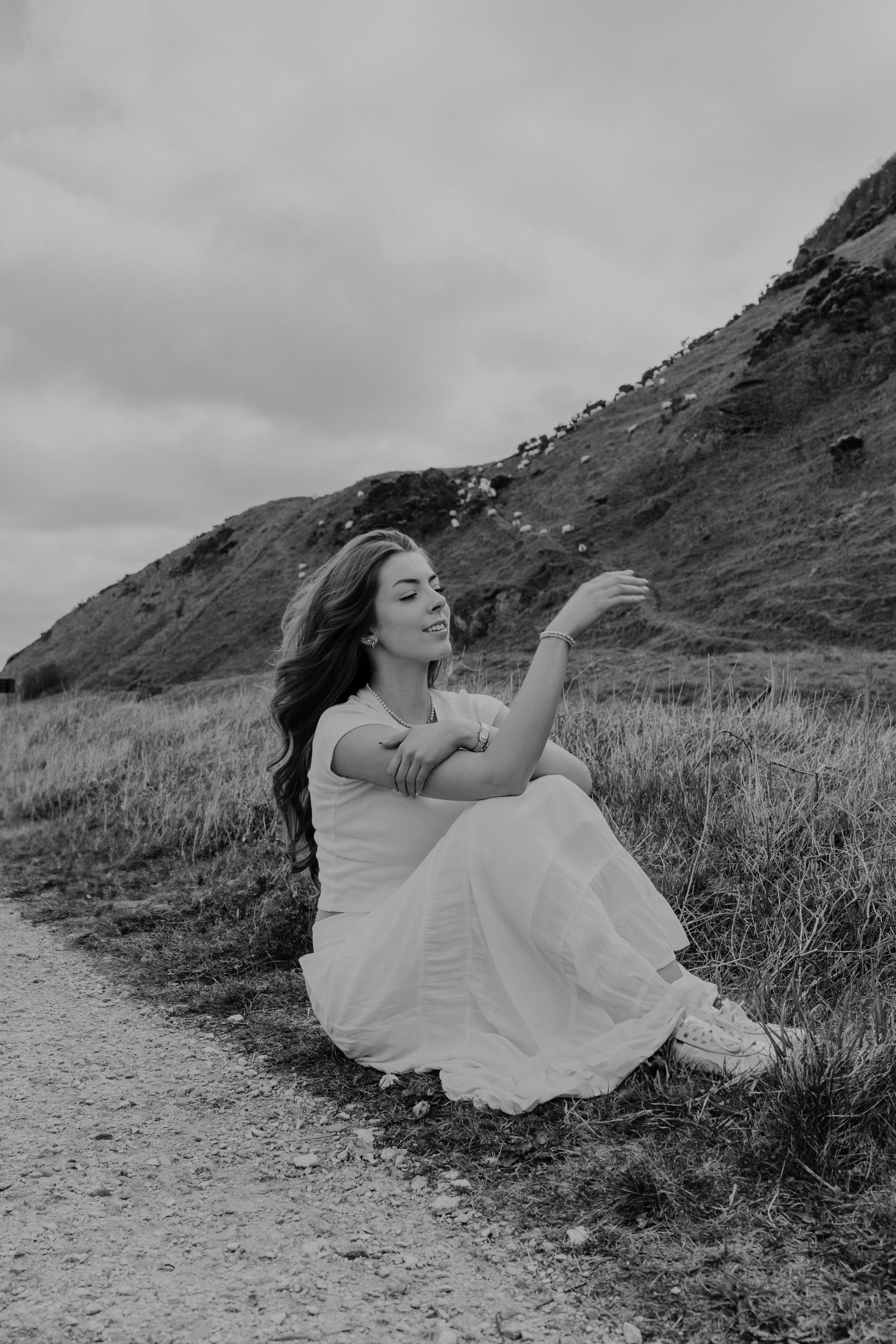 A young woman with long hair sitting on a grassy hillside, dressed in a long white skirt and t-shirt, with mountains and cloudy sky in the background.