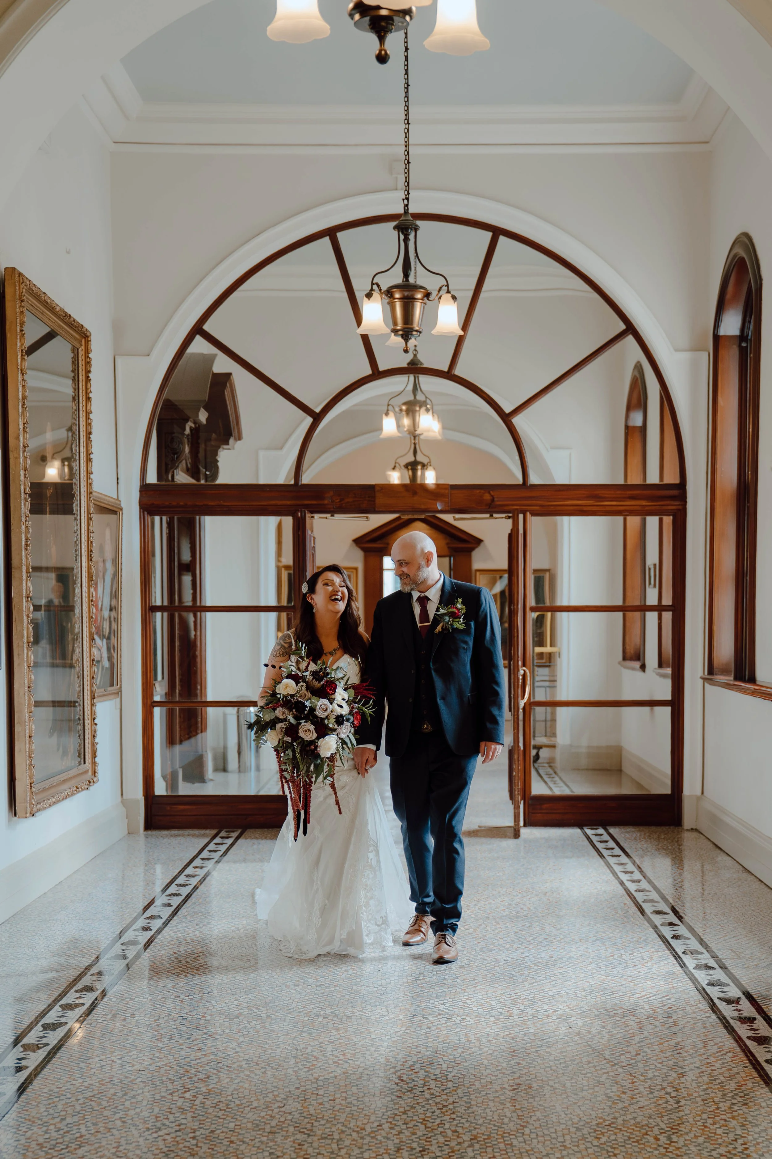 A newlywed couple walking hand in hand through an elegant hallway. The bride is holding a large bouquet and is laughing, while the groom is smiling. The hallway has arched windows, wooden accents, and vintage lighting fixtures.