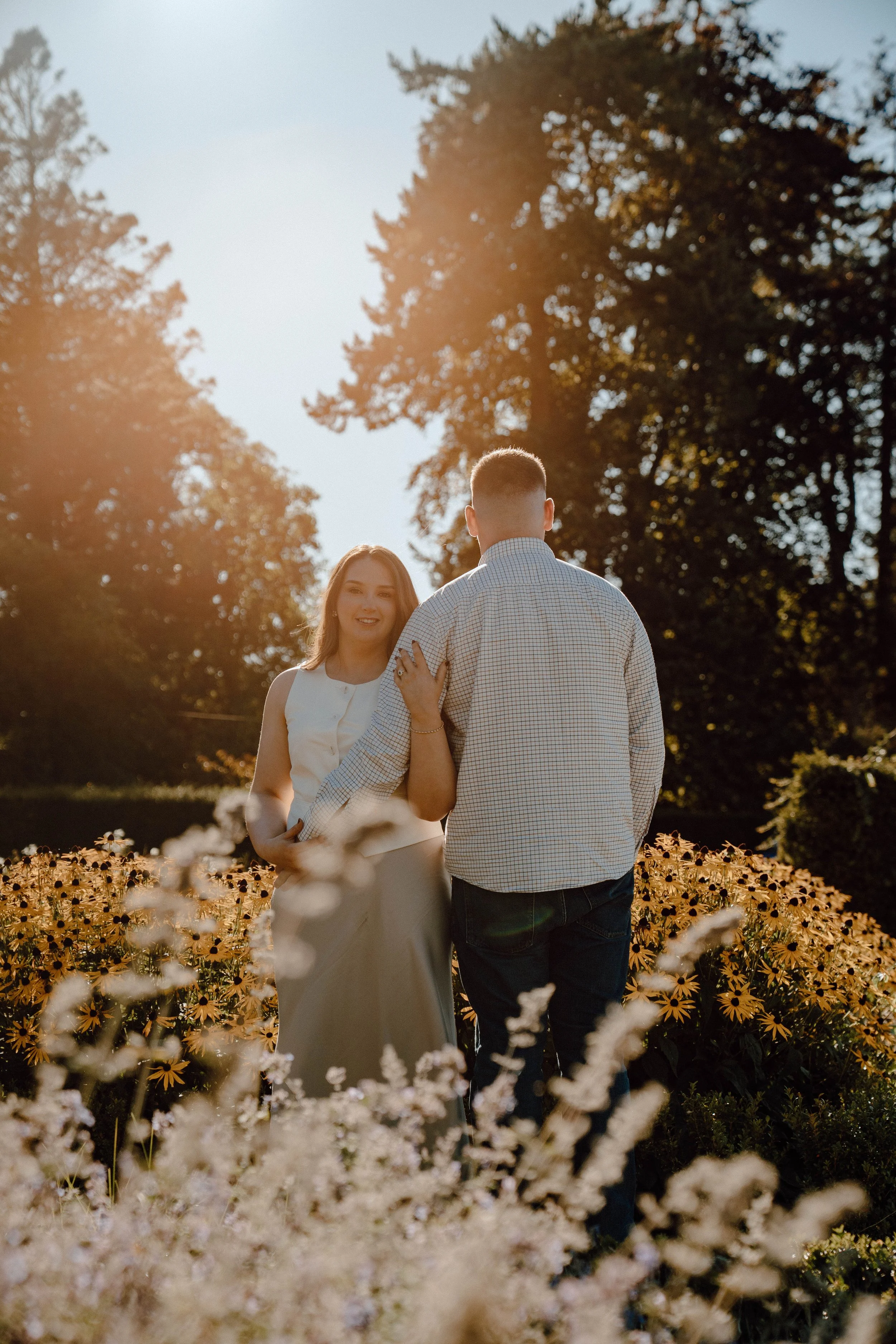 A couple stands in a flower garden during sunset, with the woman smiling as she holds the man's hand.