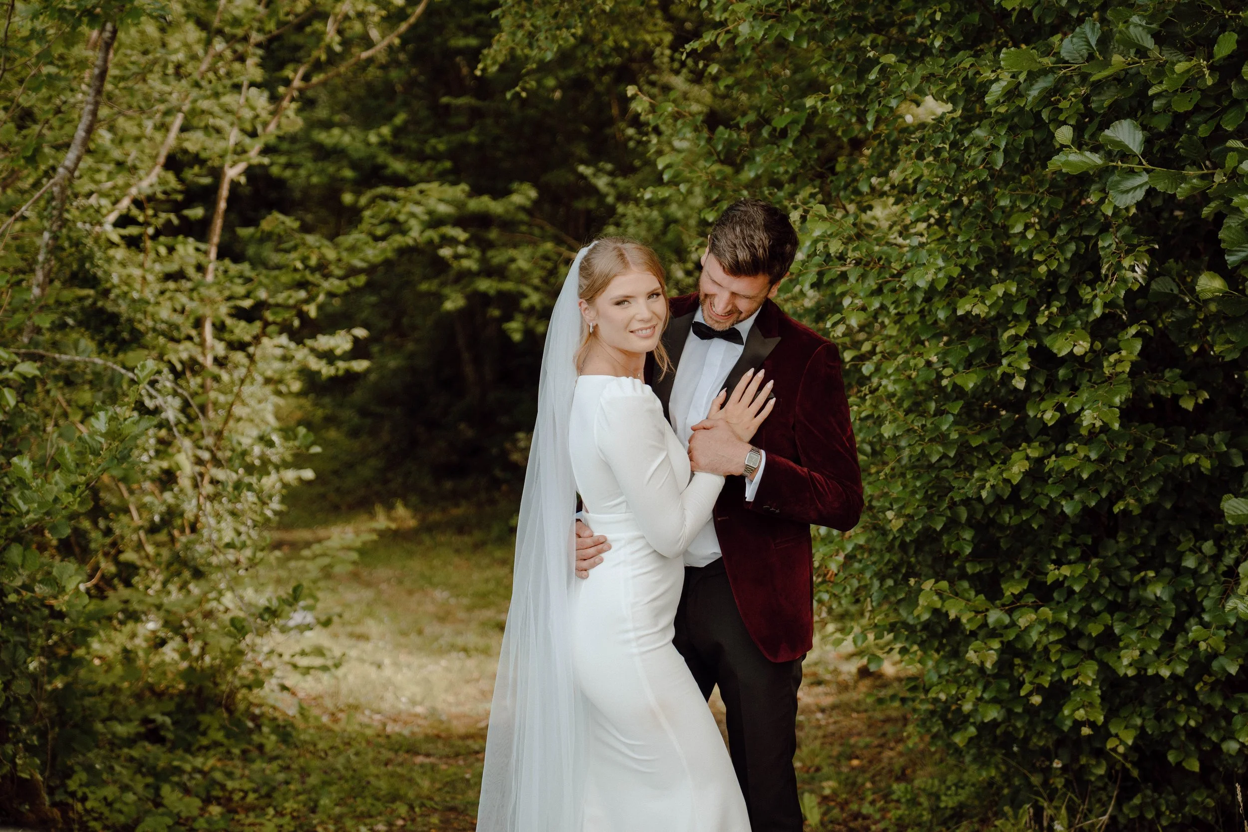 Bride and groom smiling and embracing in a wooded outdoor setting on their wedding day, with the bride wearing a white dress and veil, and the groom dressed in a tuxedo with a burgundy jacket.