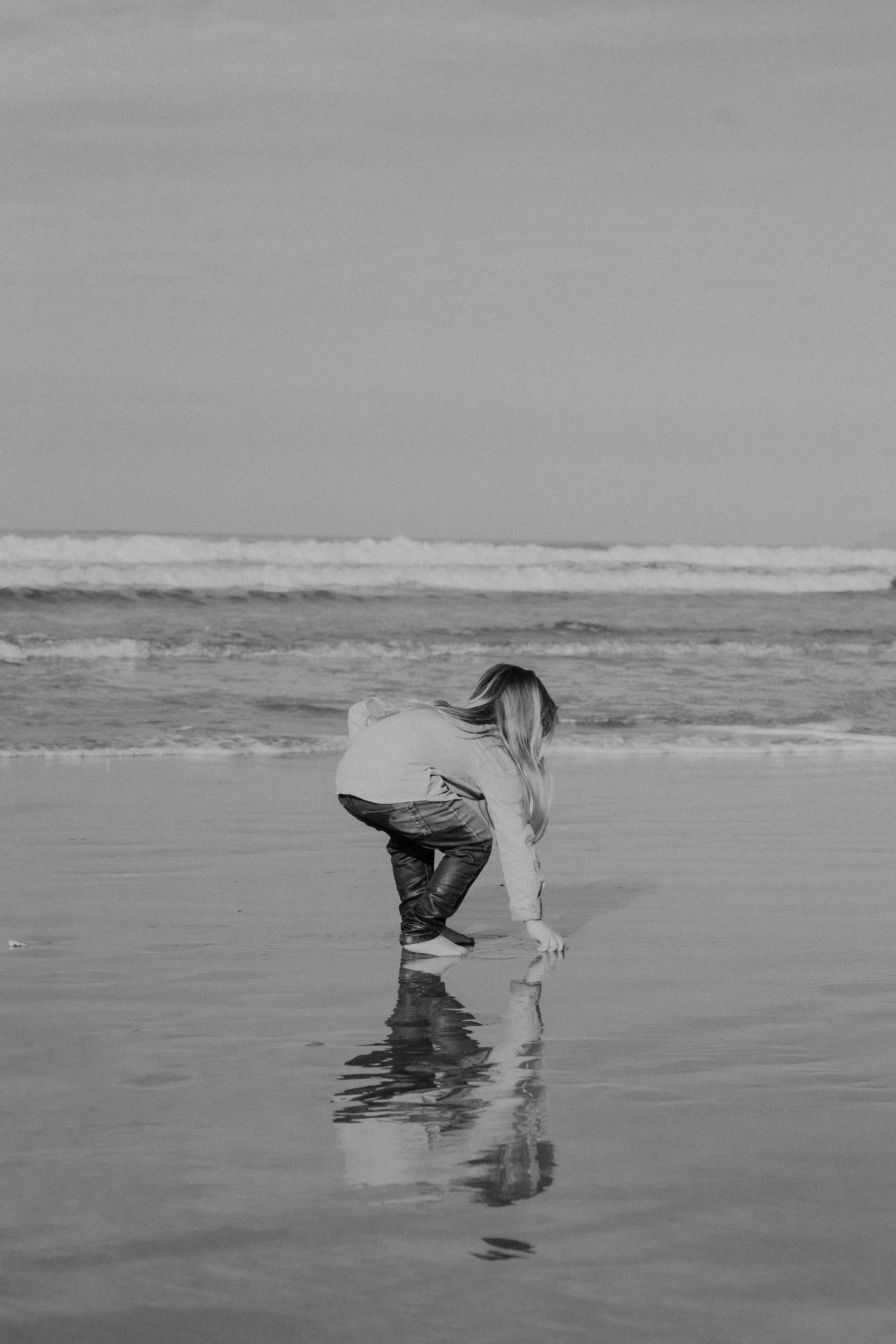 A person with long hair crouching near the shoreline, reaching into the water at a beach with gentle waves.