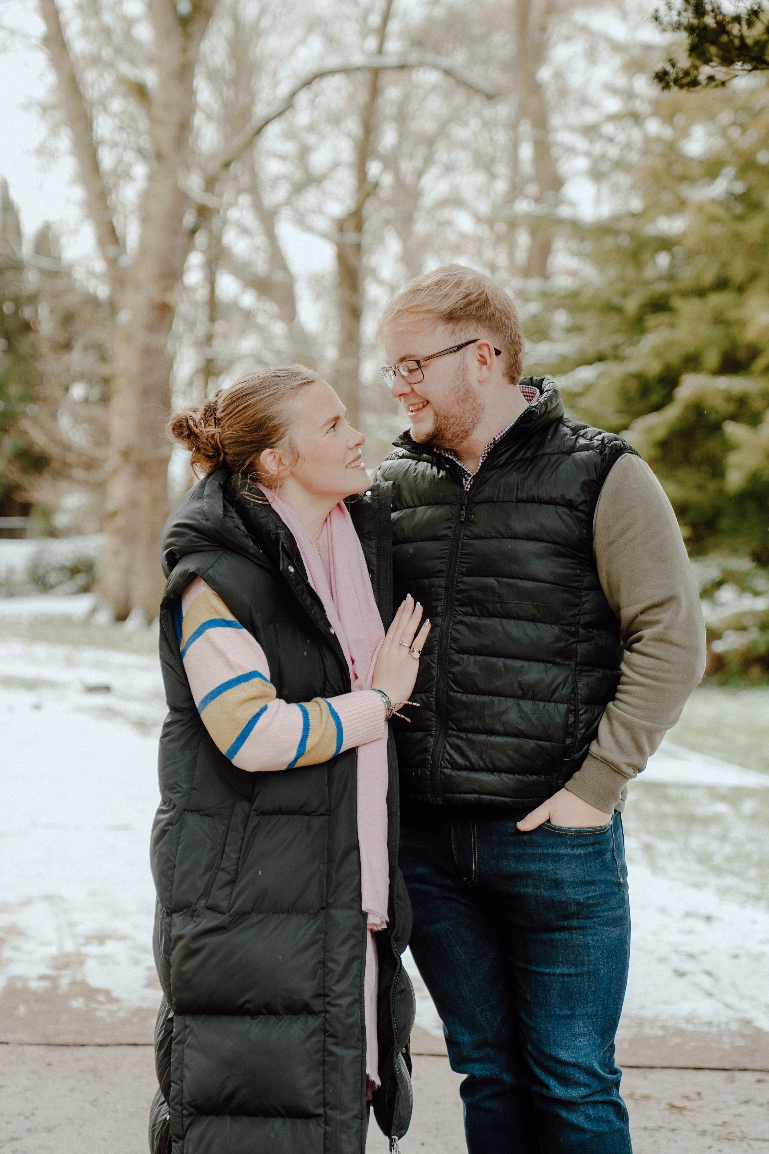 A couple in winter clothing sharing a happy moment outdoors with trees and snow in the background.