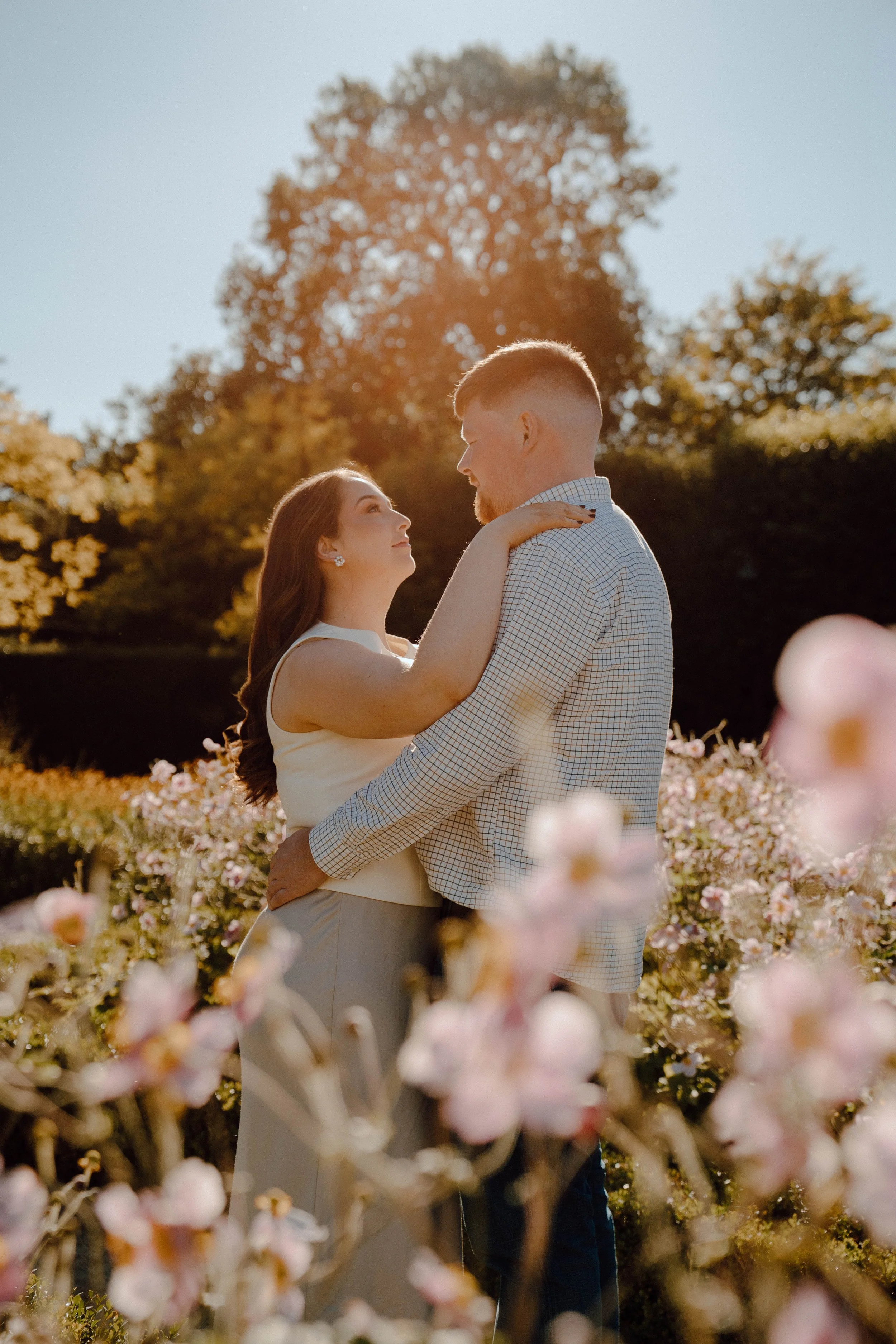 A couple stands close together outdoors among blooming flowers, gazing into each other's eyes during golden hour with trees in the background.