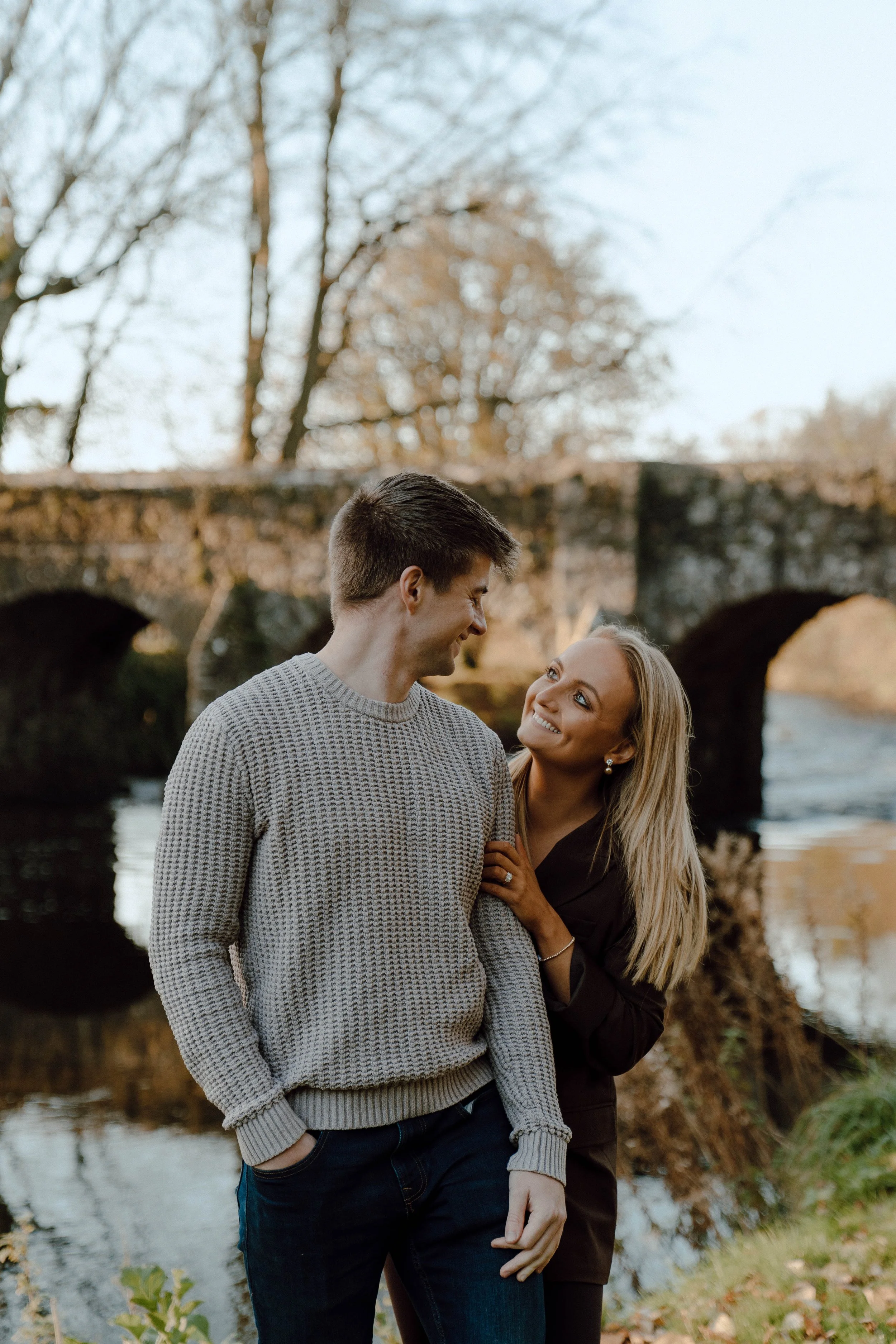 A smiling couple standing outdoors near a river with an old stone bridge in the background during autumn.
