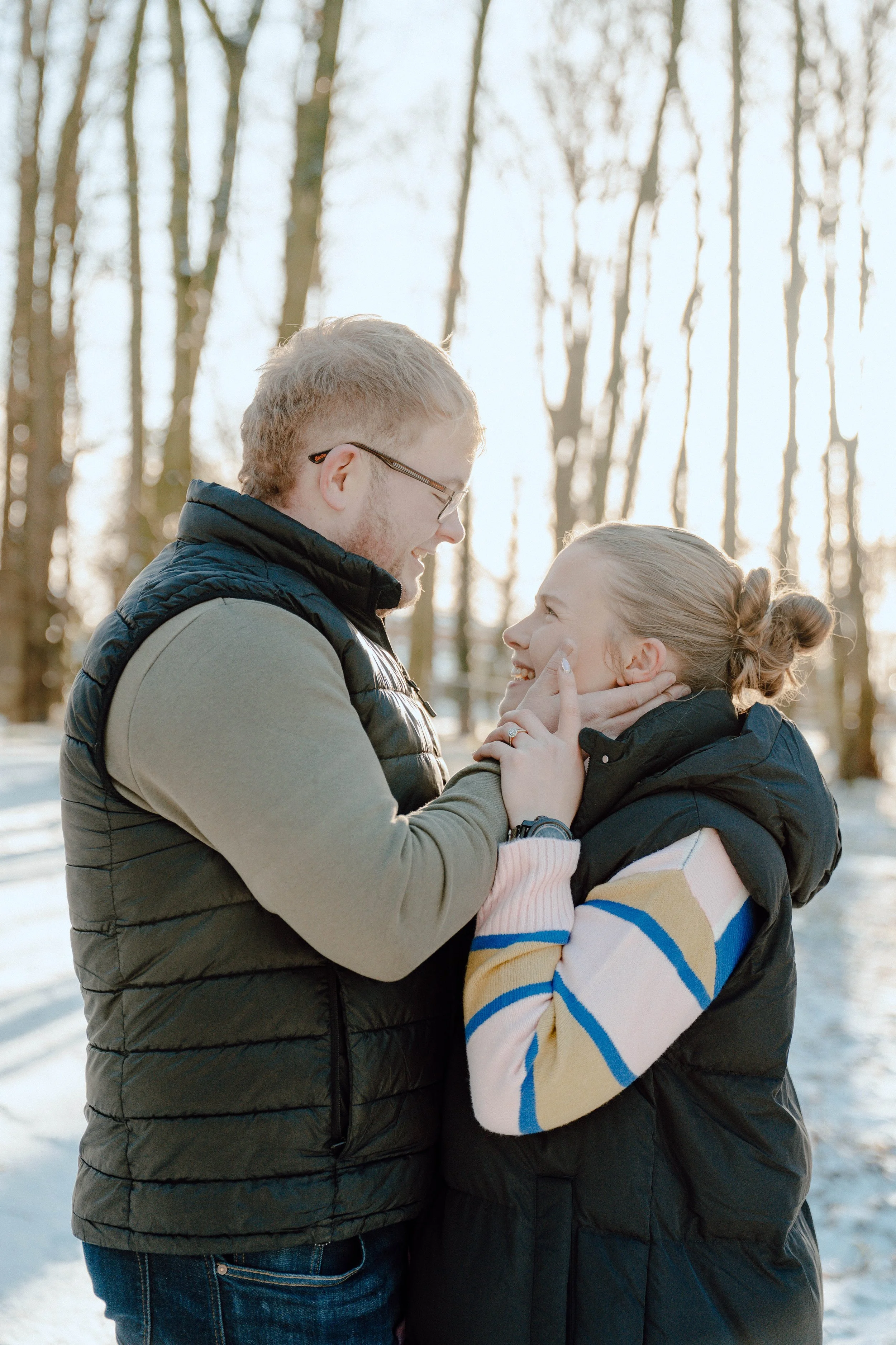 A man and woman smiling and looking at each other outdoors in a snowy forest during daytime.