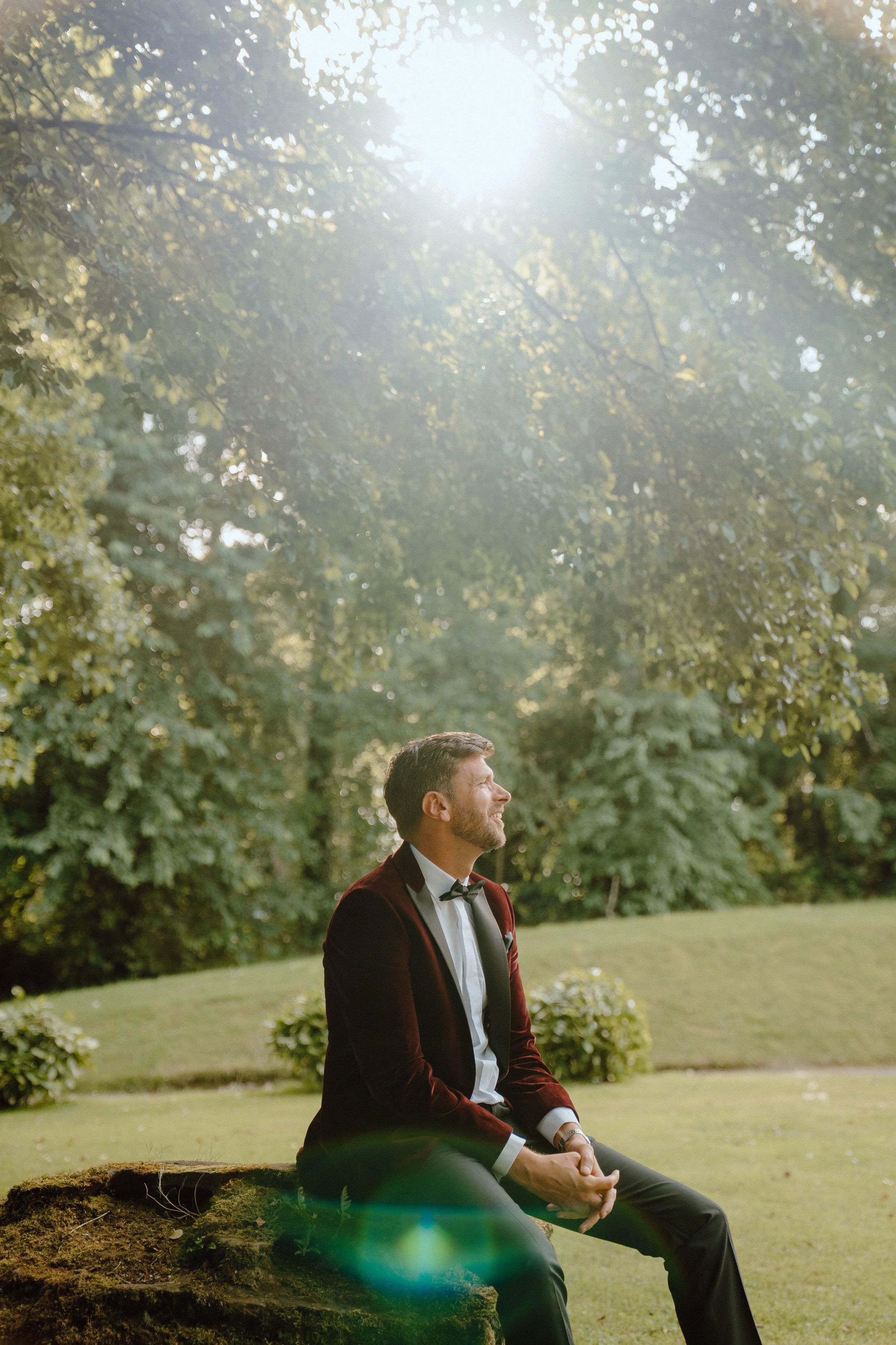 A man in a tuxedo with a bow tie sitting on a mossy rock, smiling and looking up in a park with lush green trees and bright sunlight.