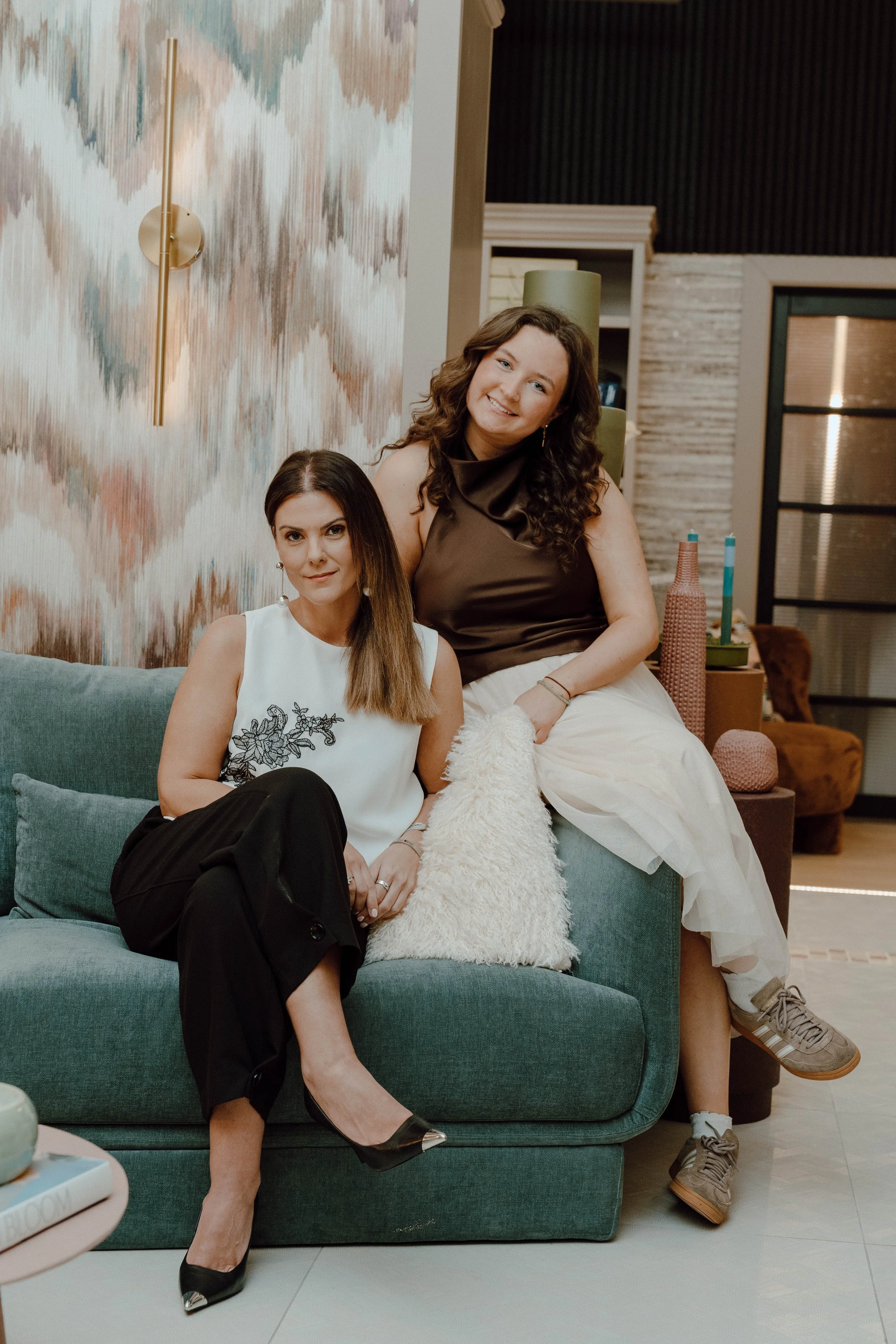 Two women sitting on a modern green sofa inside a stylish room, one sitting on the armrest and the other seated on the cushion, with decorative vases on a side table behind them.