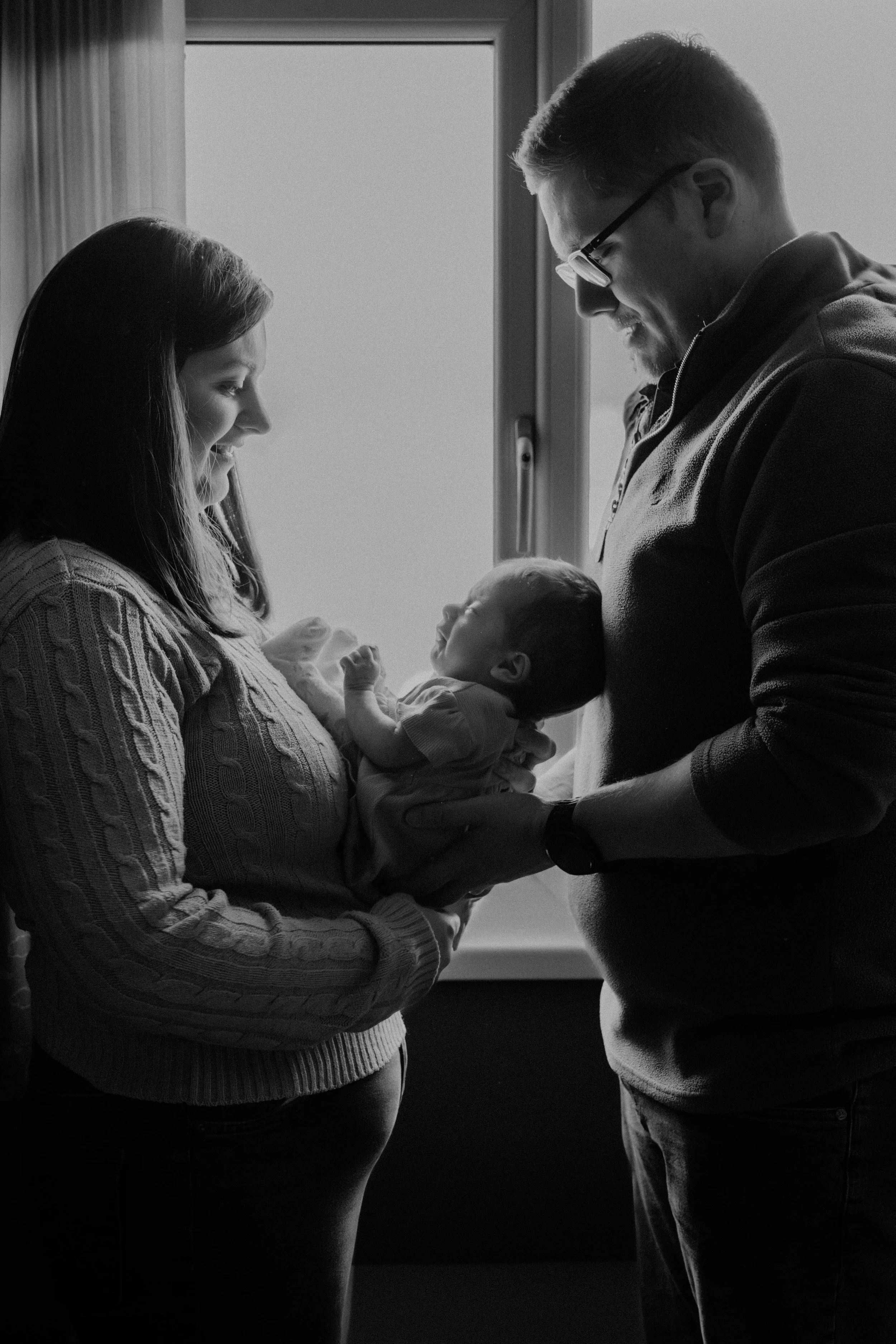 A woman holding a baby, with a man standing in front of her, all looking at each other, near a window.