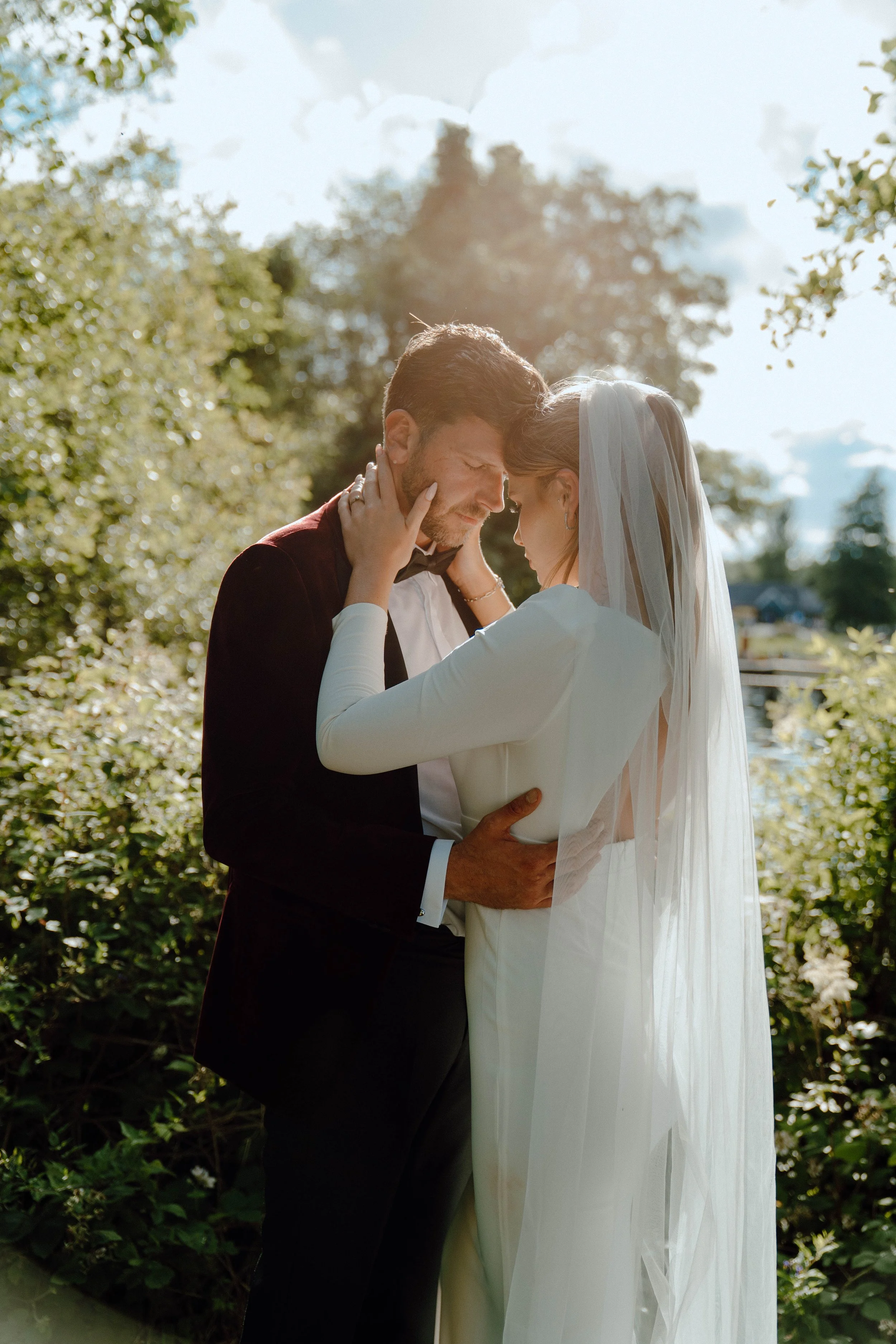 A bride and groom are holding each other closely outdoors during their wedding, with trees and a river in the background, as they share an intimate moment, touching foreheads.